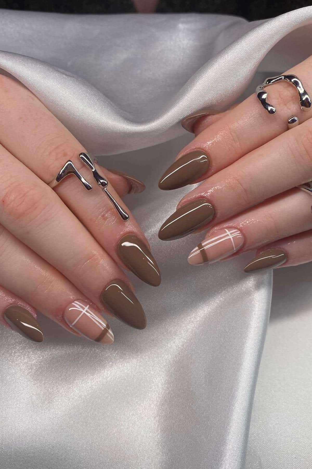 Hands with long, almond-shaped nails painted in brown and nude tones, featuring plaid accent designs, resting on a white satin fabric. Silver rings are worn on several fingers.