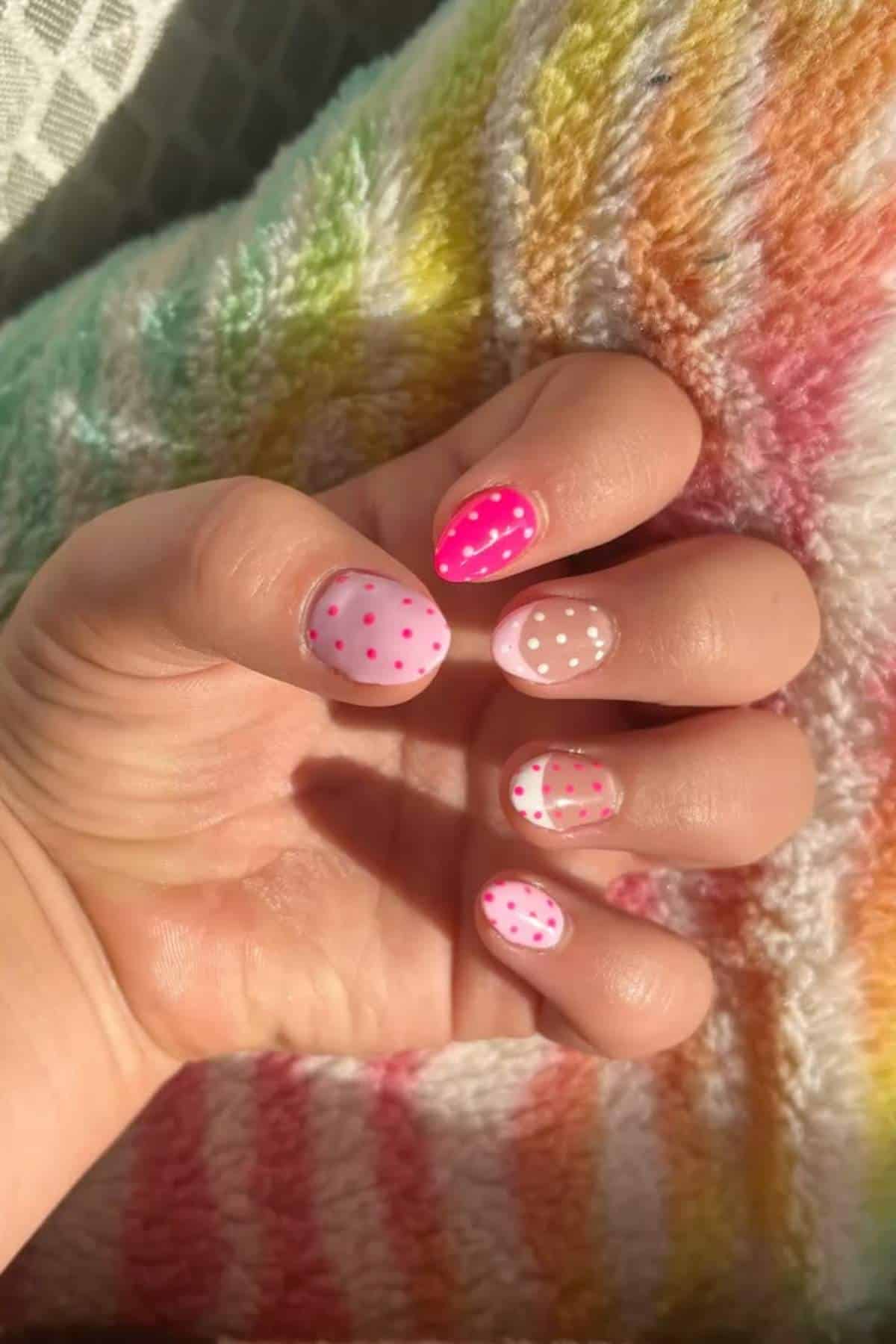 A hand with short nails painted in pink and white shades with polka dot patterns, resting on a multicolored, striped fabric.