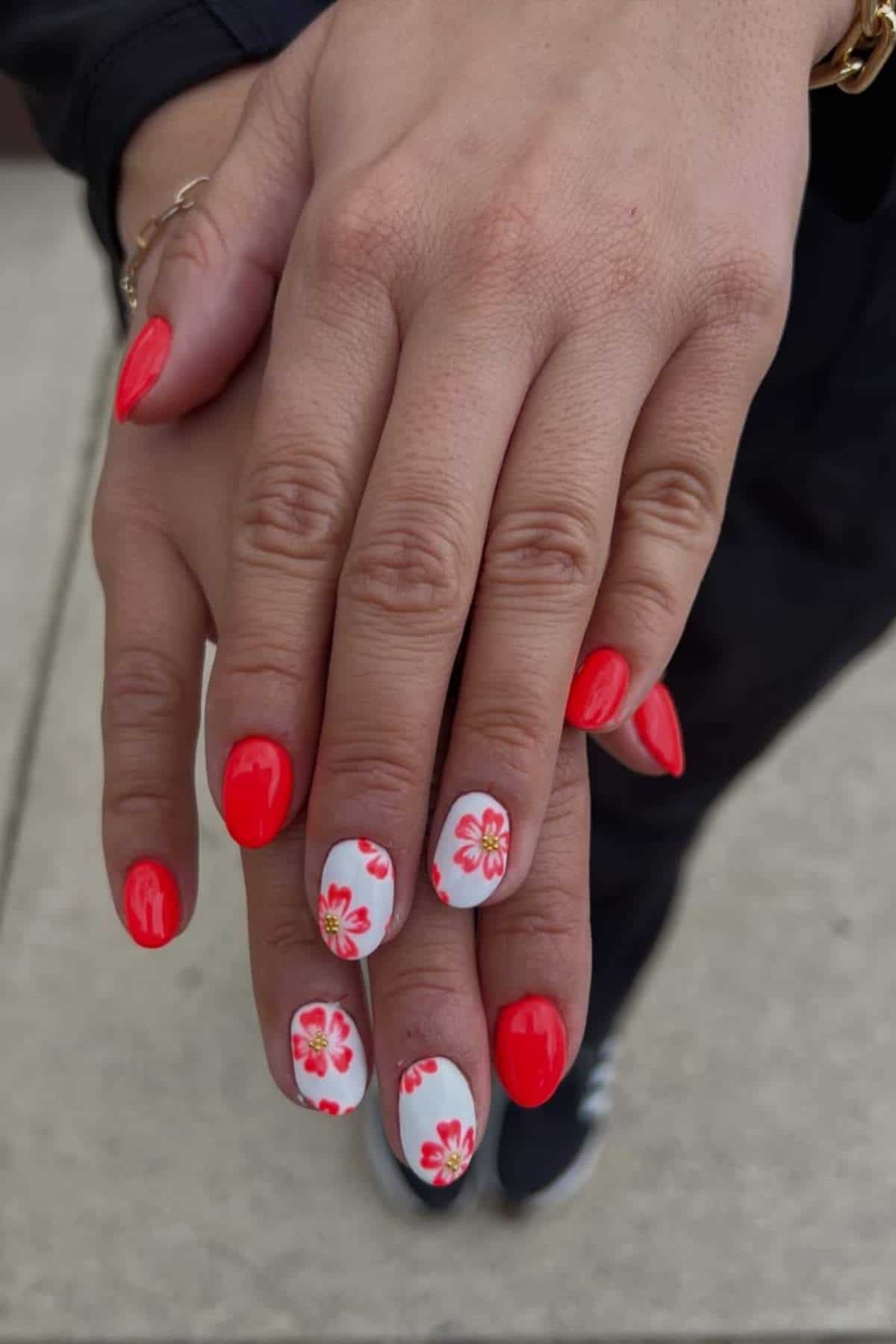 A person with red and white floral nail art rests one hand on top of the other, displaying manicured nails against a blurred outdoor background.