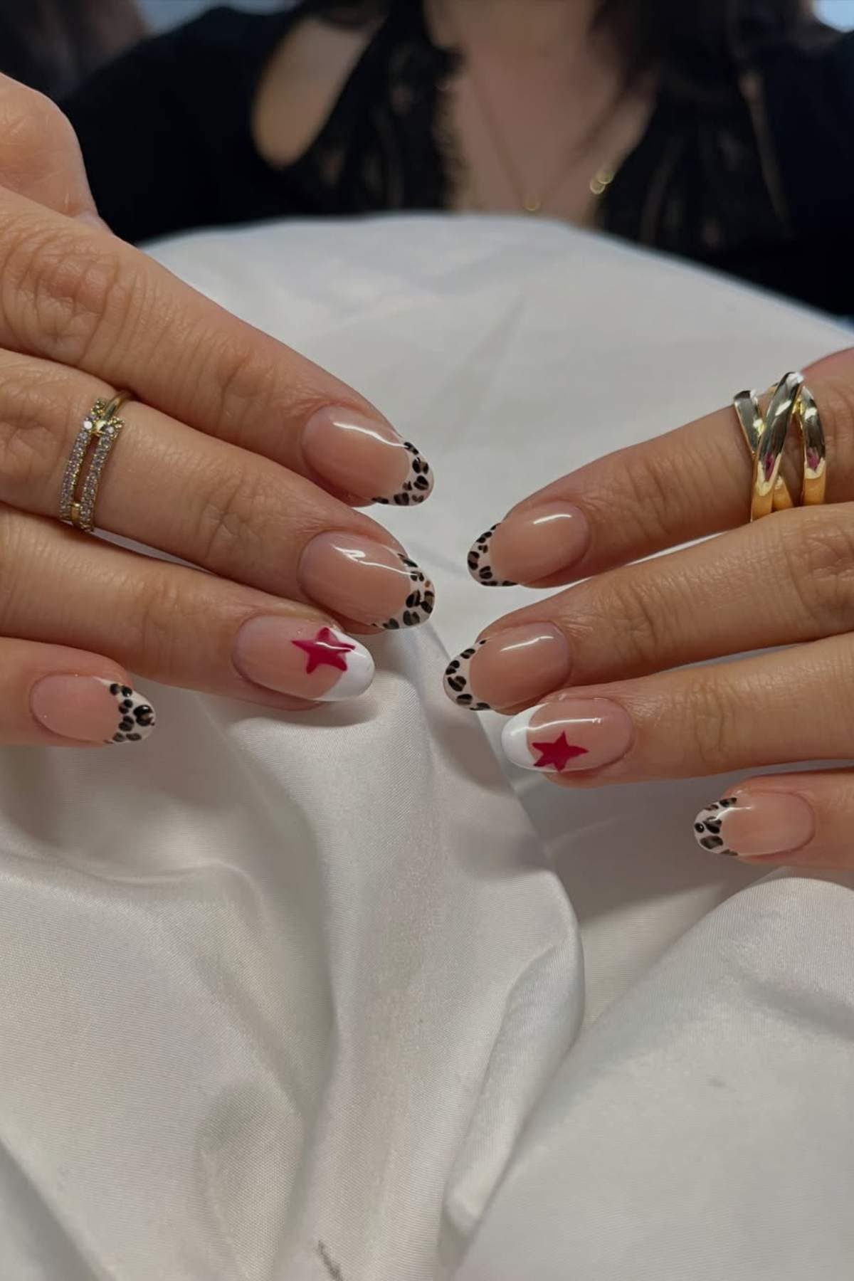 Hands with manicured nails featuring leopard print French tips and two accent nails with white tips and red stars, resting on a white fabric.