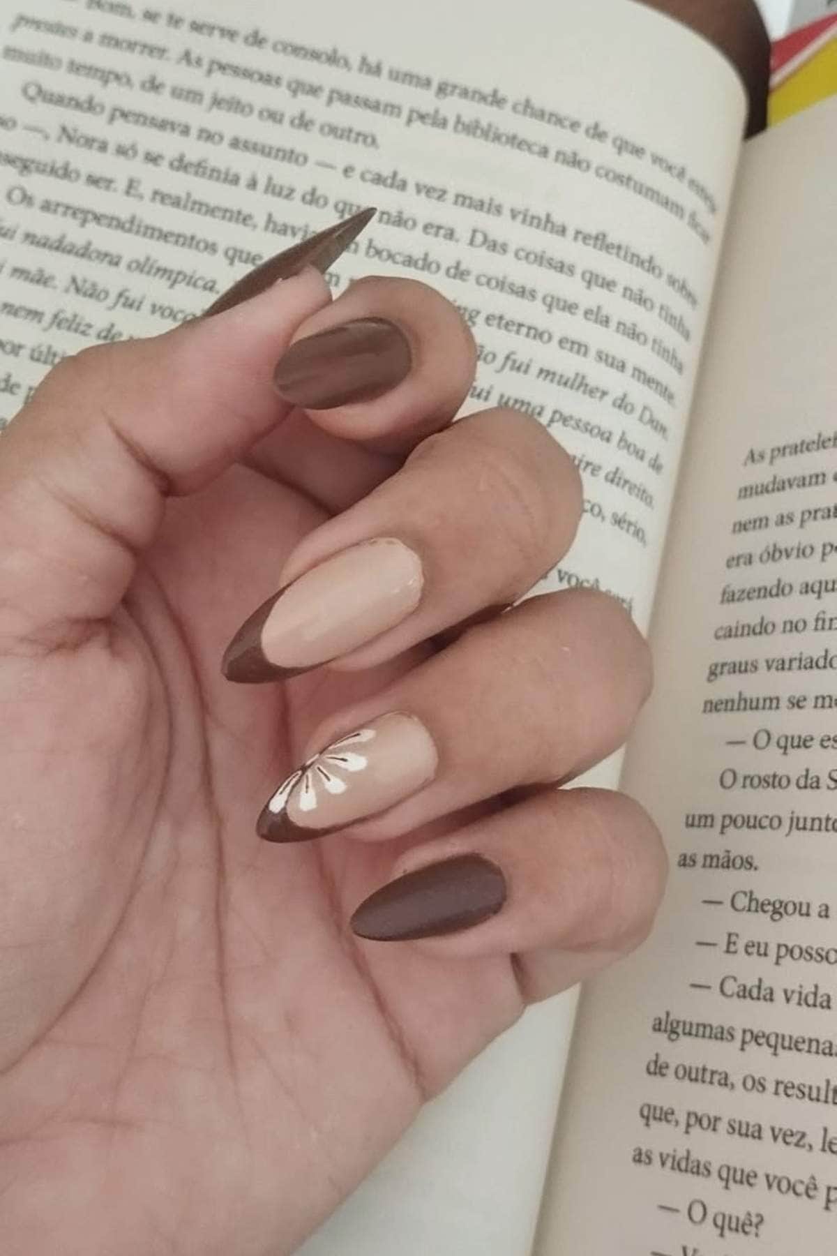 A hand with almond-shaped nails painted in matte brown and beige tones, featuring a white leaf design on the ring fingernail, is holding an open book.