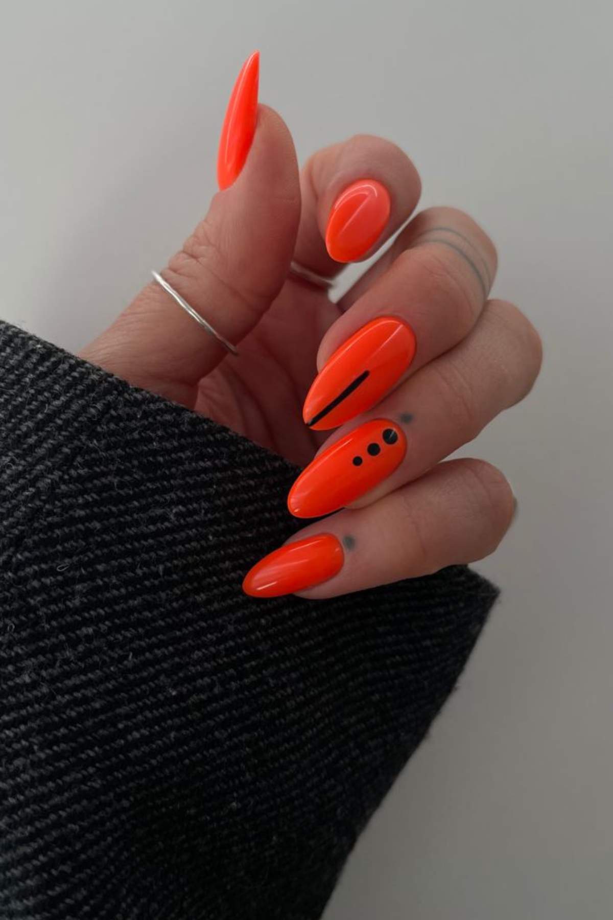 A hand with long, almond-shaped neon orange nails; one nail has a thin black line and another has three black dots. The person wears a dark textured sleeve and silver rings.