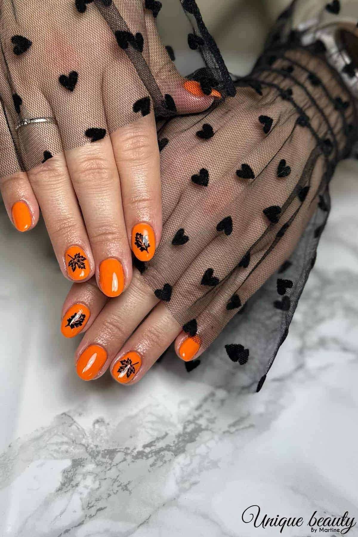 Hands with bright orange nail polish, some nails feature black leaf designs. The person is wearing a sheer black mesh sleeve with heart patterns.