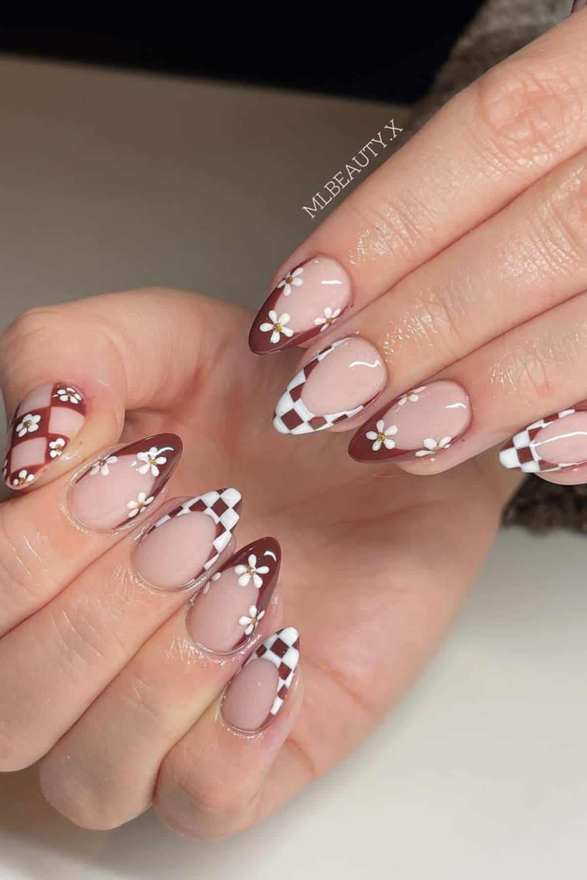 Hands with almond-shaped nails featuring brown and white checkerboard patterns, French tips, and small white daisy flower designs.