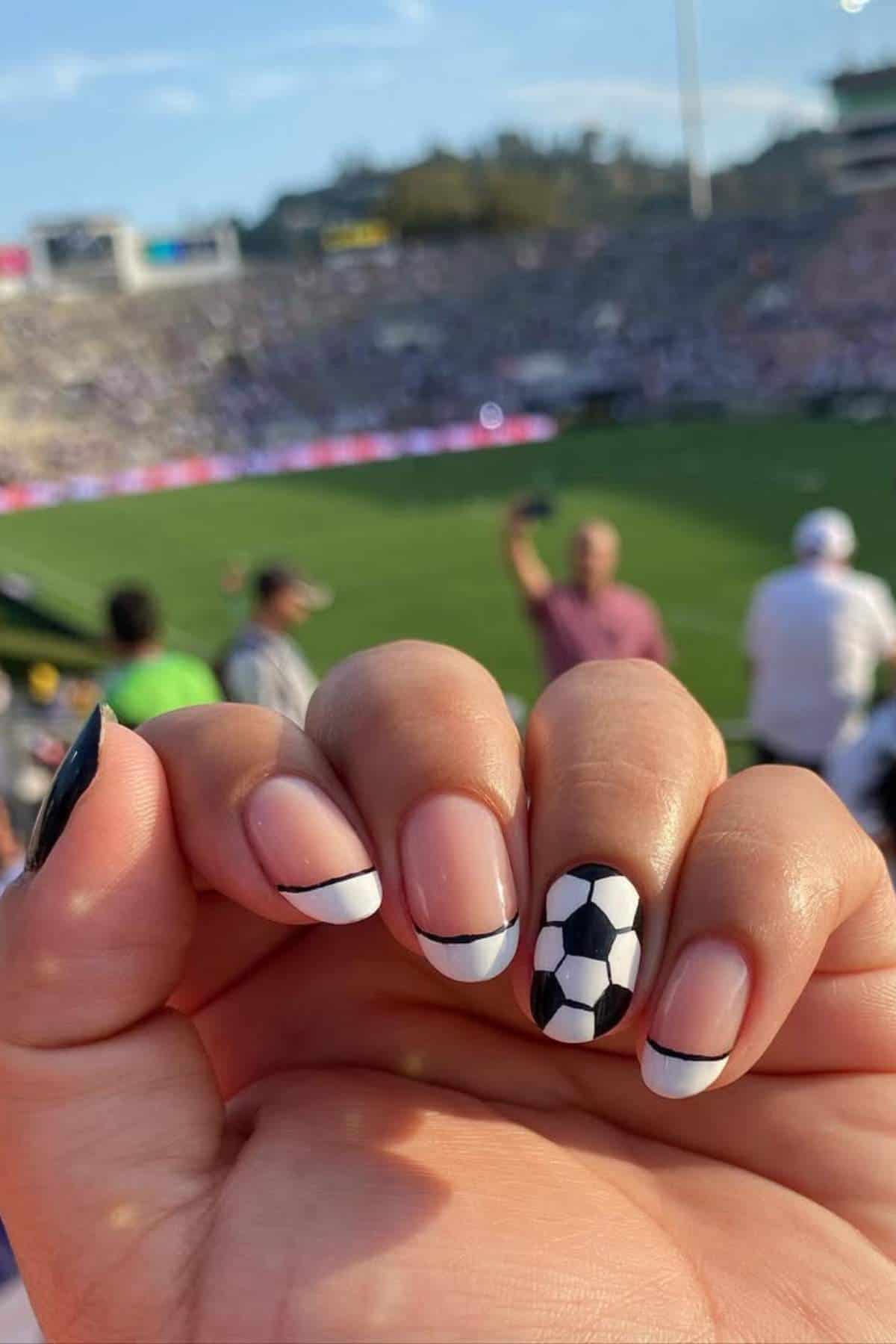A hand with manicured nails, one featuring a soccer ball design, is held up at a soccer stadium with a blurred crowd and field in the background.