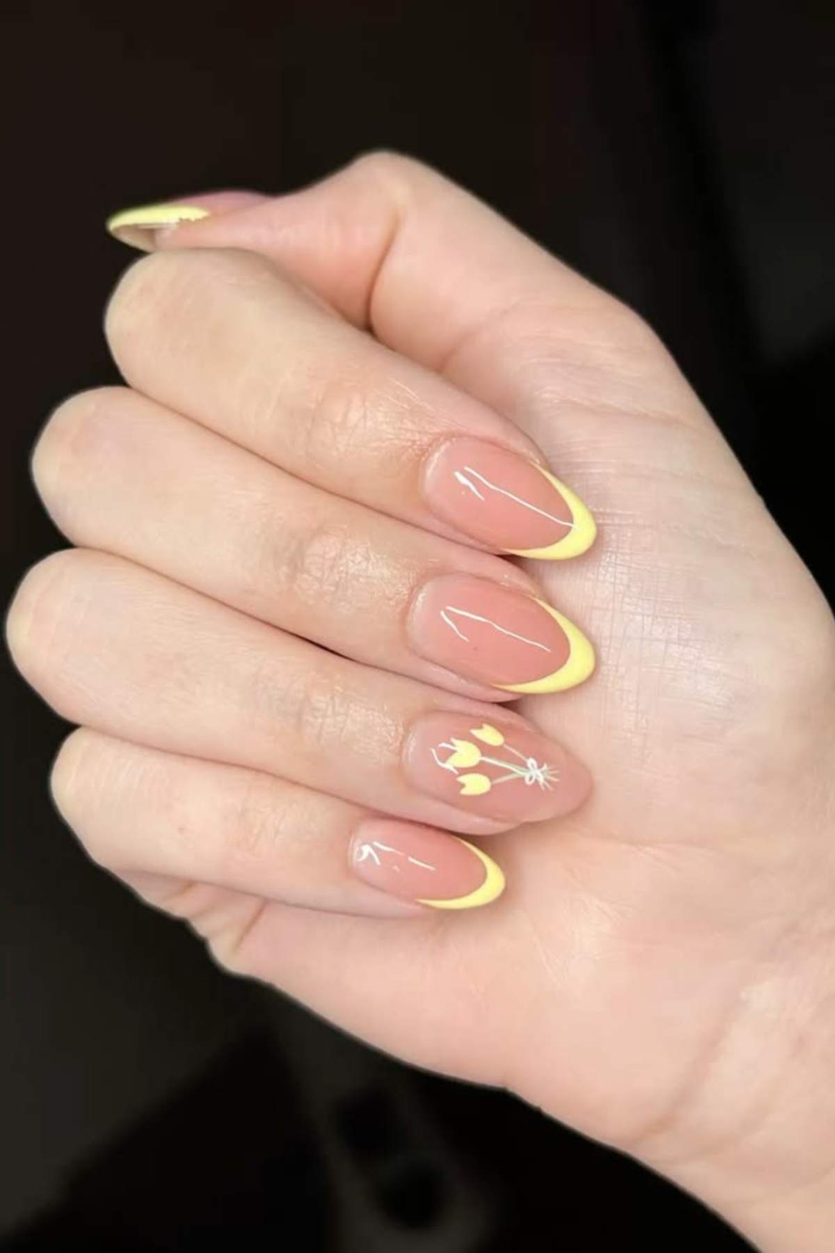 A hand with almond-shaped nails featuring yellow French tips and white floral designs on two nails.