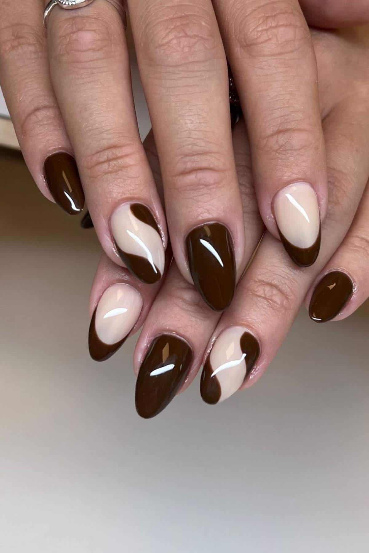 Hands with almond-shaped nails painted in a brown and beige abstract pattern, alternating between solid and two-tone designs.