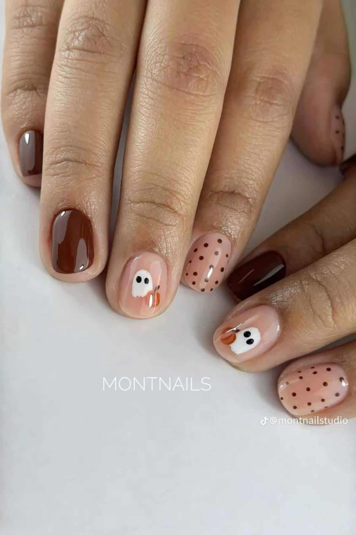 A hand with short nails painted in brown and nude tones, featuring polka dots and simple turkey faces as Thanksgiving-themed nail art.