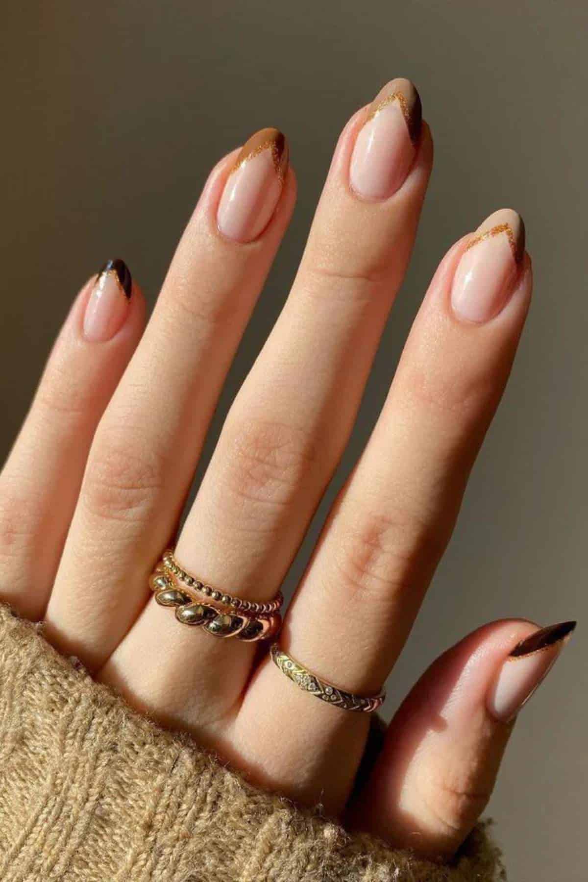 A hand with almond-shaped nails featuring brown and gold French tips, wearing two rings, and partially covered by a beige knit sleeve.
