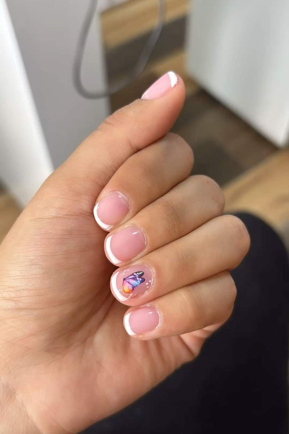 A hand with short, natural nails featuring a French manicure and a butterfly design on the ring fingernail.