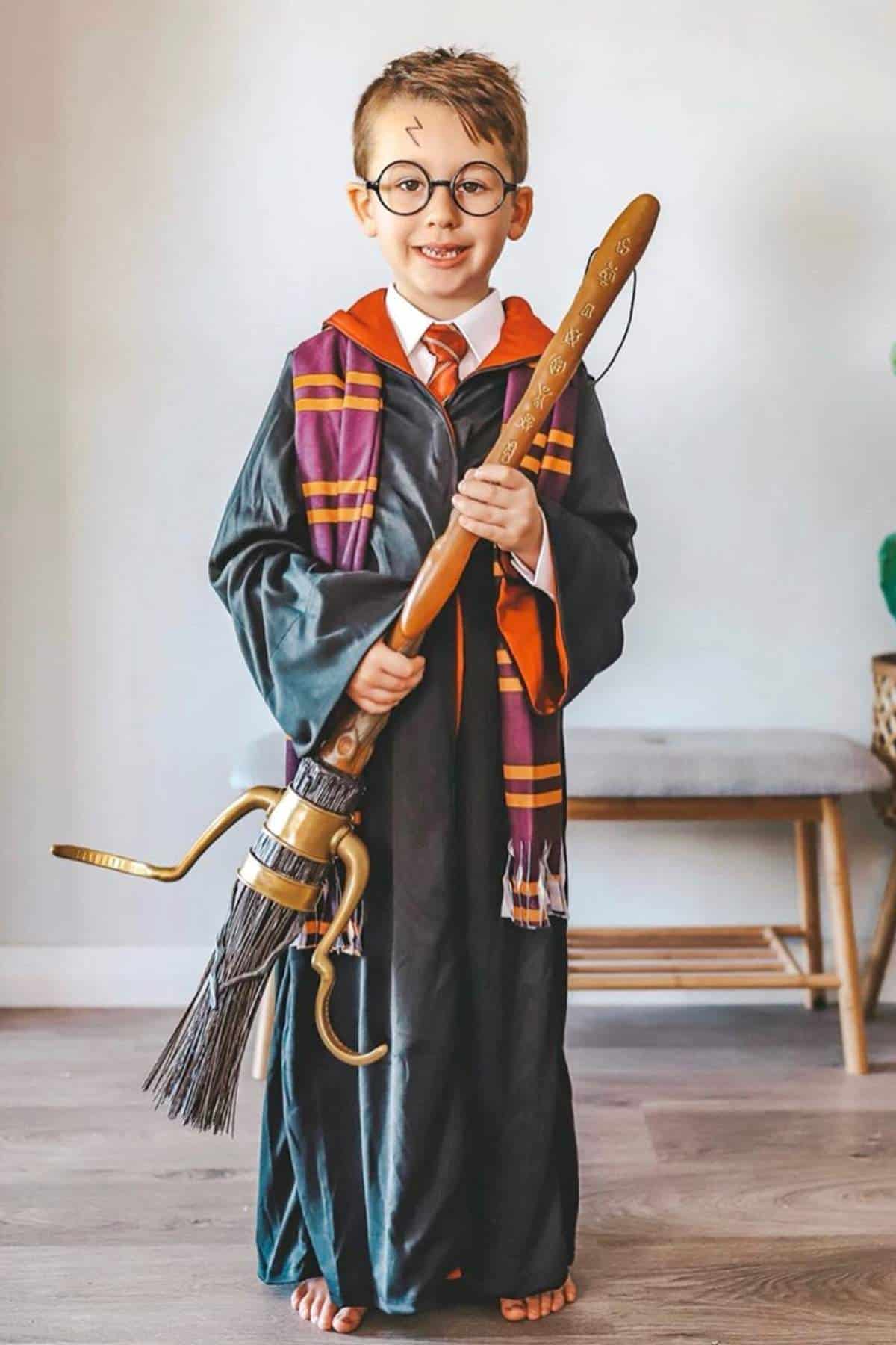 A young child in a Harry Potter costume holds a broomstick while smiling at the camera, standing barefoot on a wooden floor.