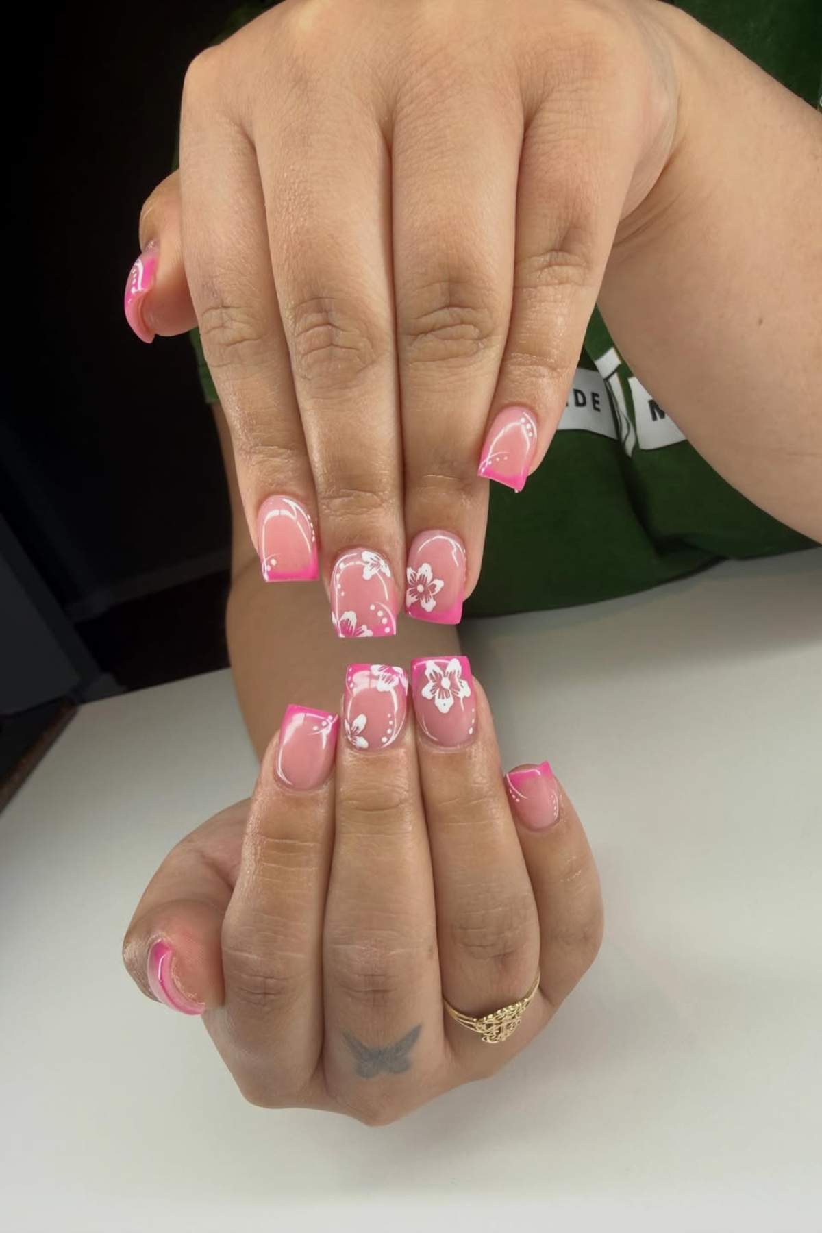 Two hands with square-shaped nails featuring a pink French tip and white floral designs on a few nails, resting on a white surface.