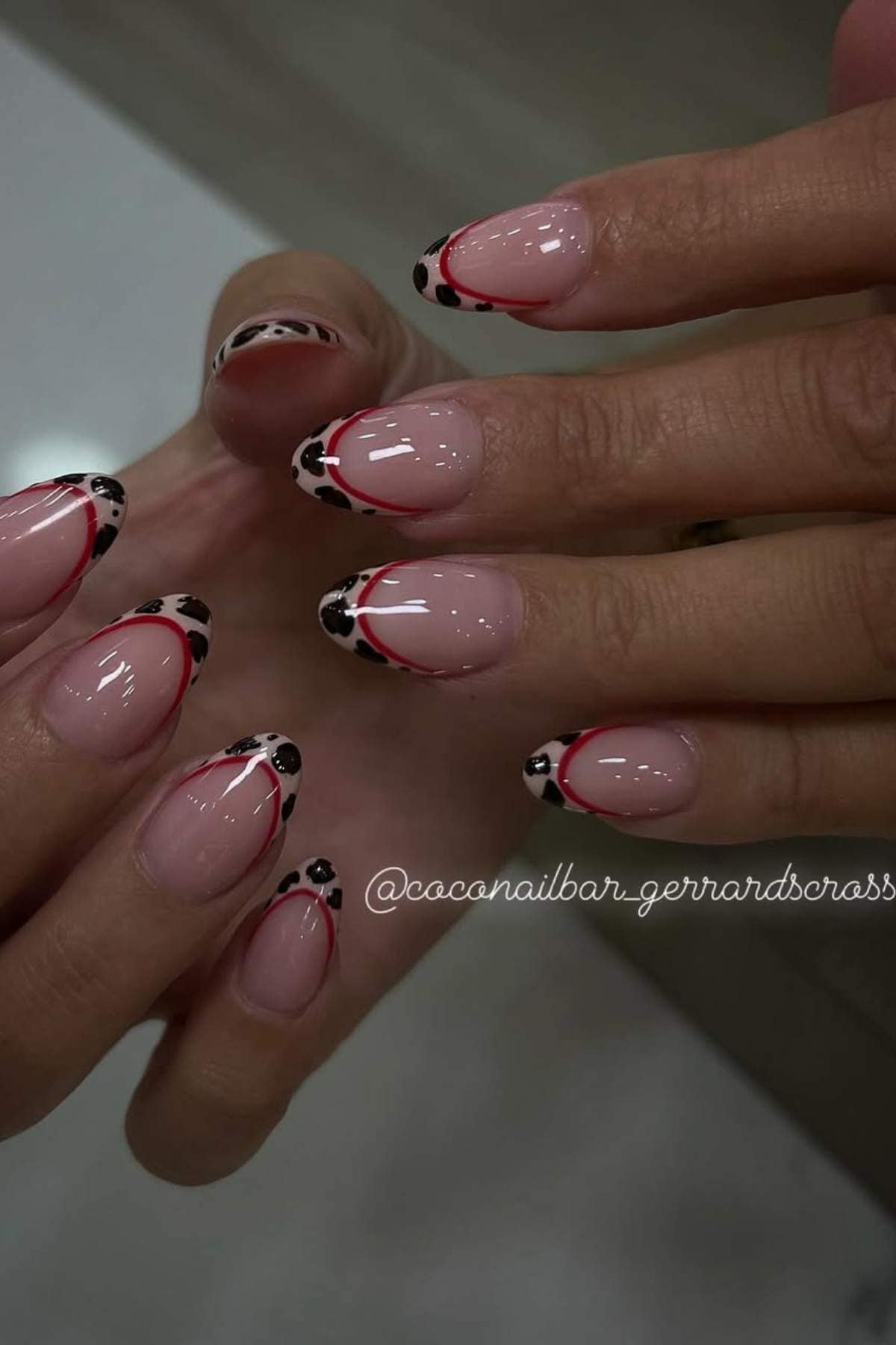 Close-up of hands with almond-shaped nails featuring a nude base, red and black abstract French tips, and black-and-white spotted accents.