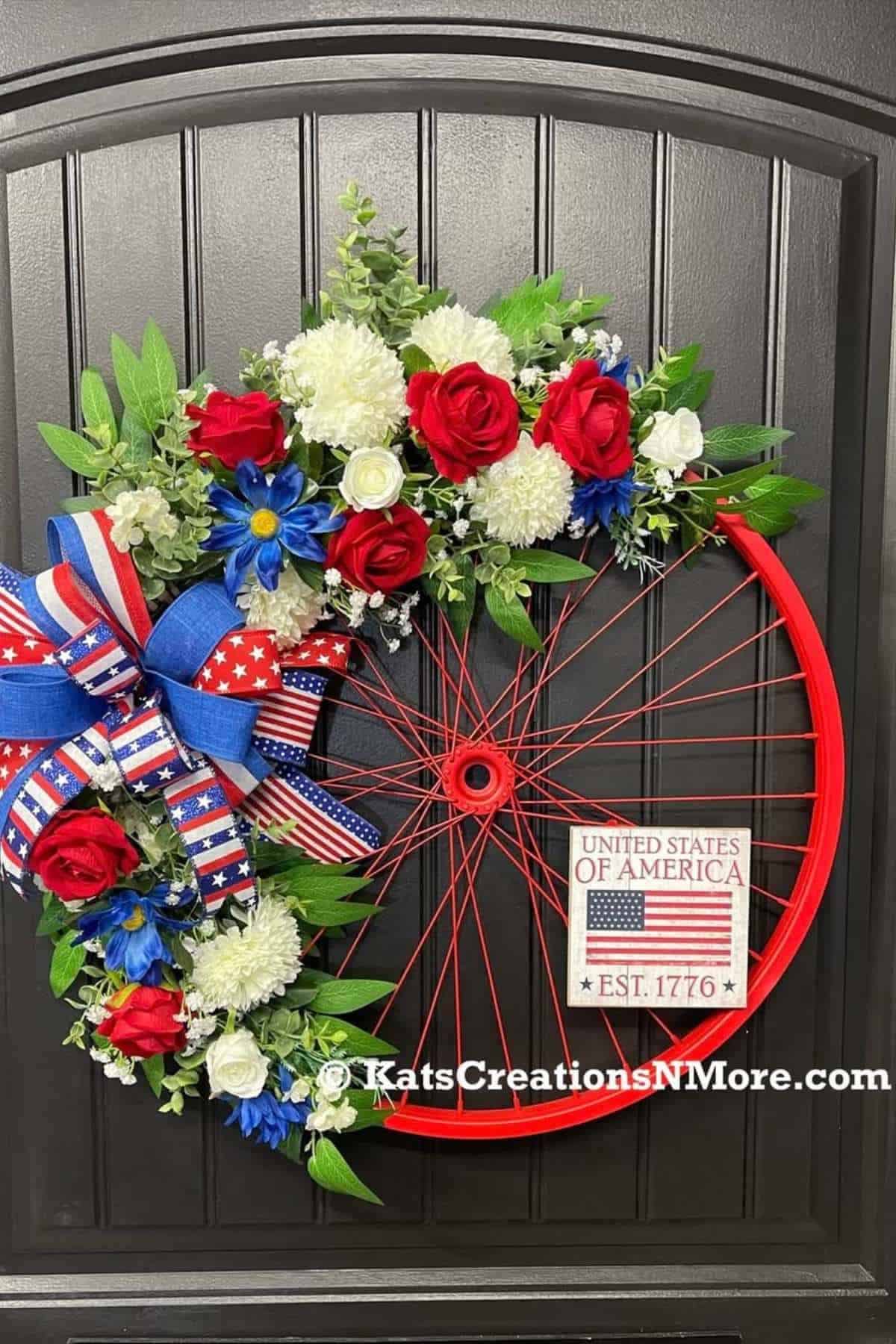 A red bicycle wheel wreath decorated with red, white, and blue flowers, patriotic ribbons, and a sign that reads "United States of America Est. 1776" on a black door.