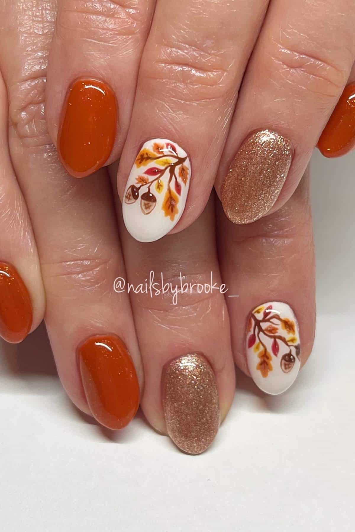 Close-up of hands with autumn-themed manicure featuring burnt orange, gold glitter, and white nails with fall leaf and acorn designs.