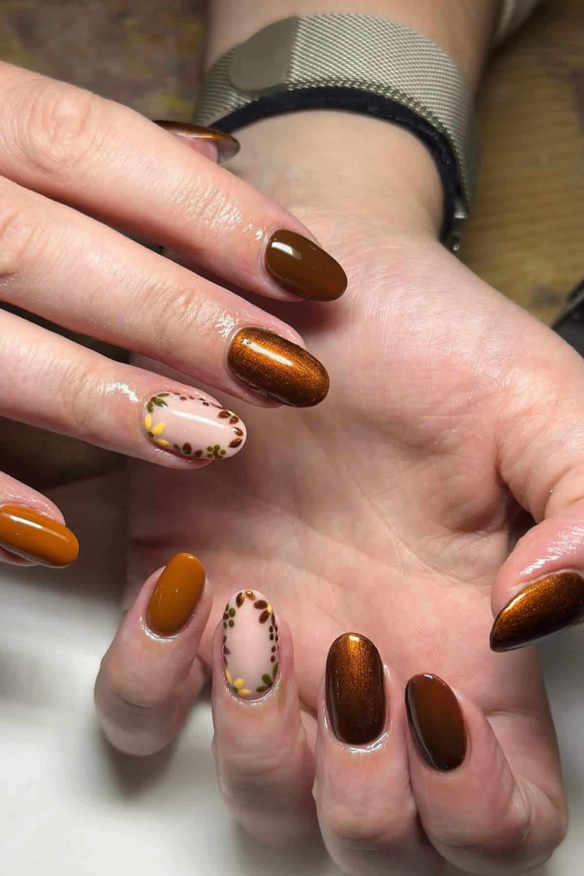 Hands with almond-shaped nails painted in brown and copper tones; two accent nails feature a nude base with a floral and dot design.