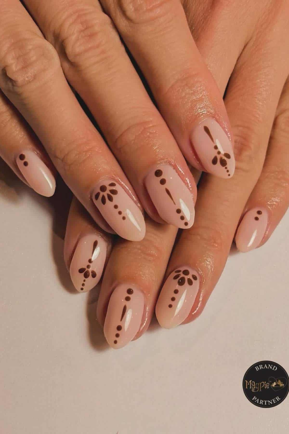 A hand with light pink nails featuring simple brown dot and line designs, some with small petal-like patterns near the cuticles.