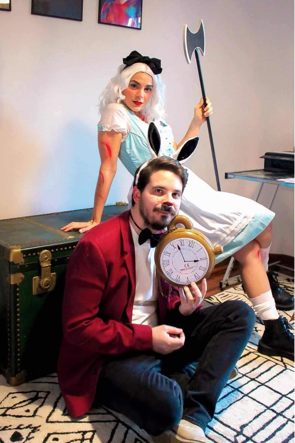 Two people in costumes; one as Alice with a blue dress, white apron, and axe, the other as the White Rabbit with bunny ears, red jacket, and large pocket watch, sitting on the floor.