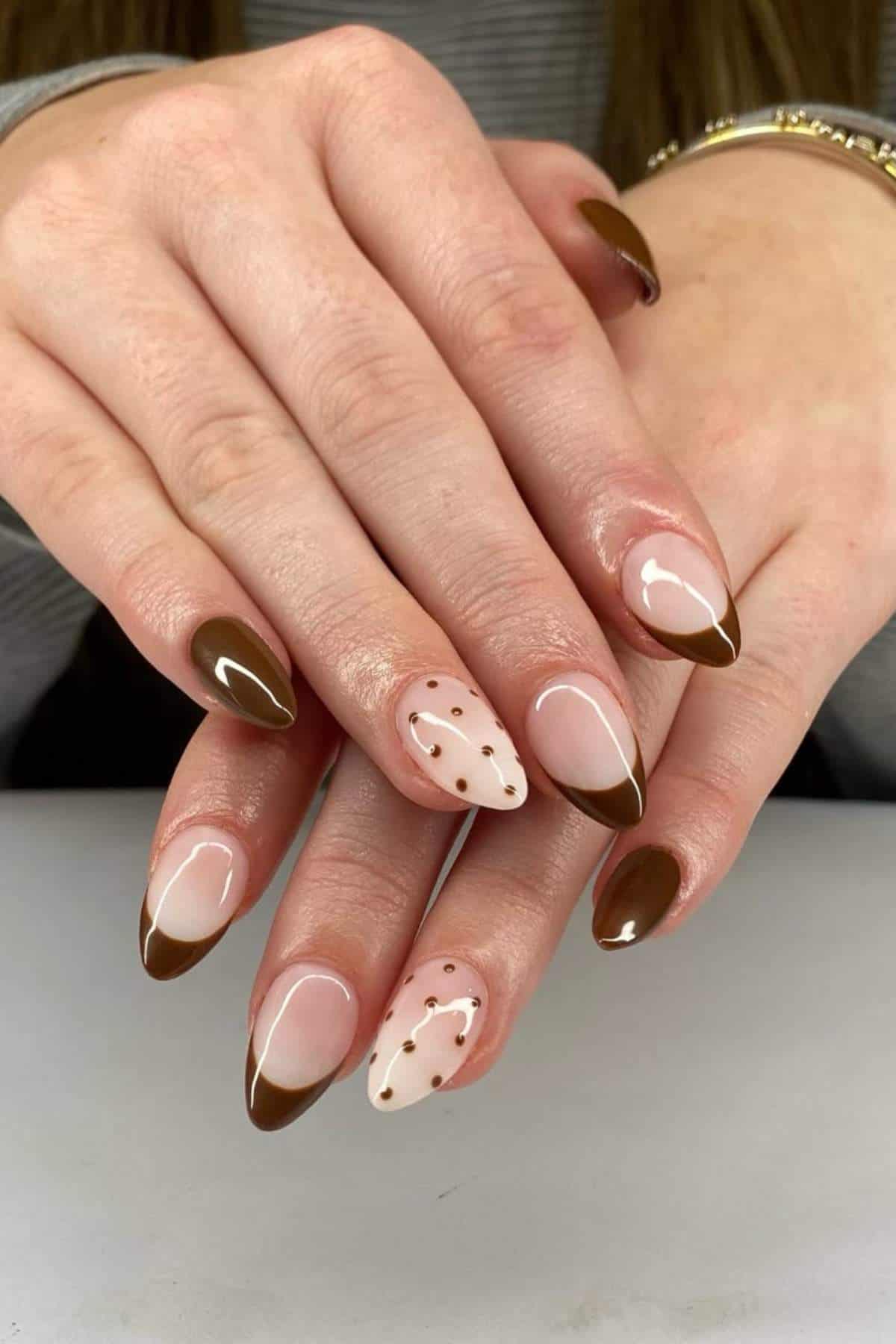Hands with almond-shaped nails painted in brown and nude pink, featuring brown French tips and polka dot accents on two nails.