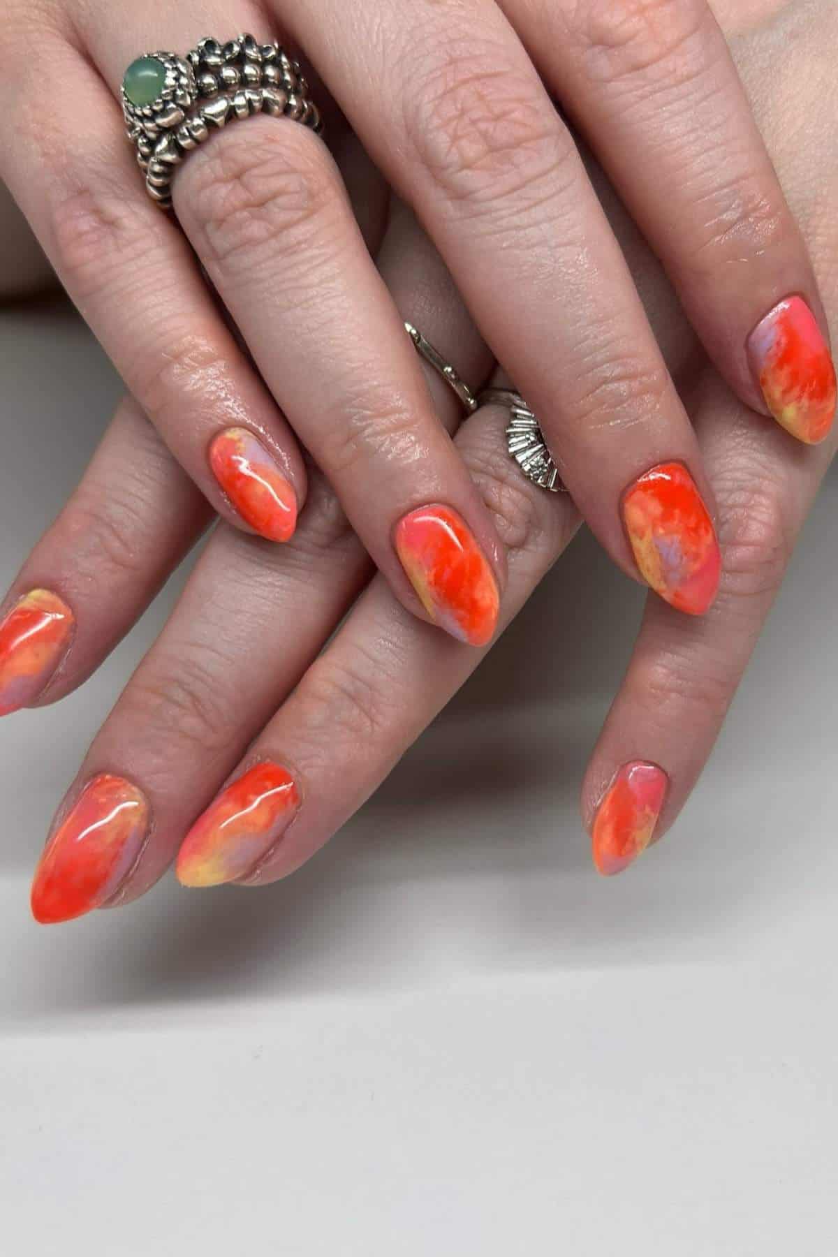 Hands with orange and yellow marble-patterned almond-shaped nails; fingers adorned with silver rings, shown against a plain background.