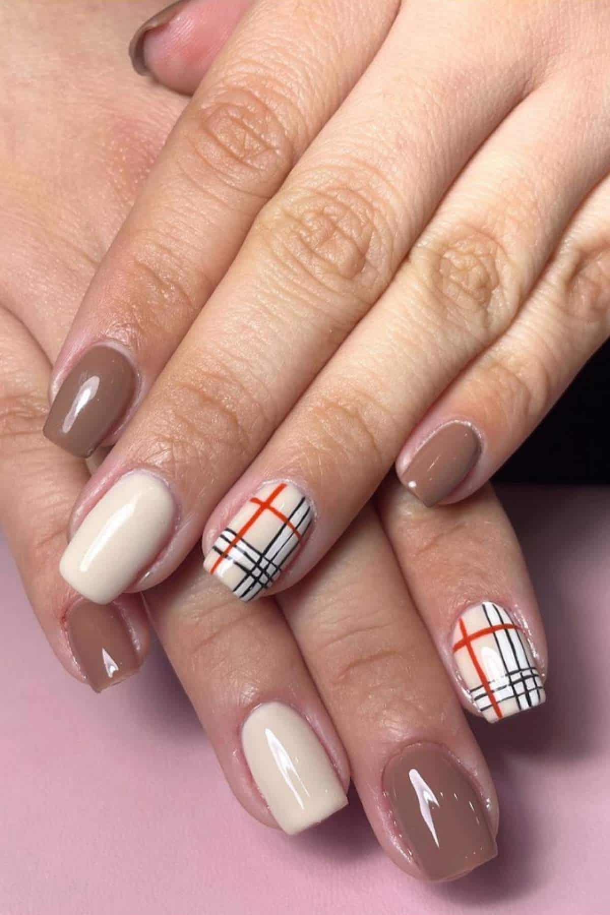 Hands with manicured nails featuring beige and brown polish; two accent nails have a plaid pattern with black, white, and orange lines.
