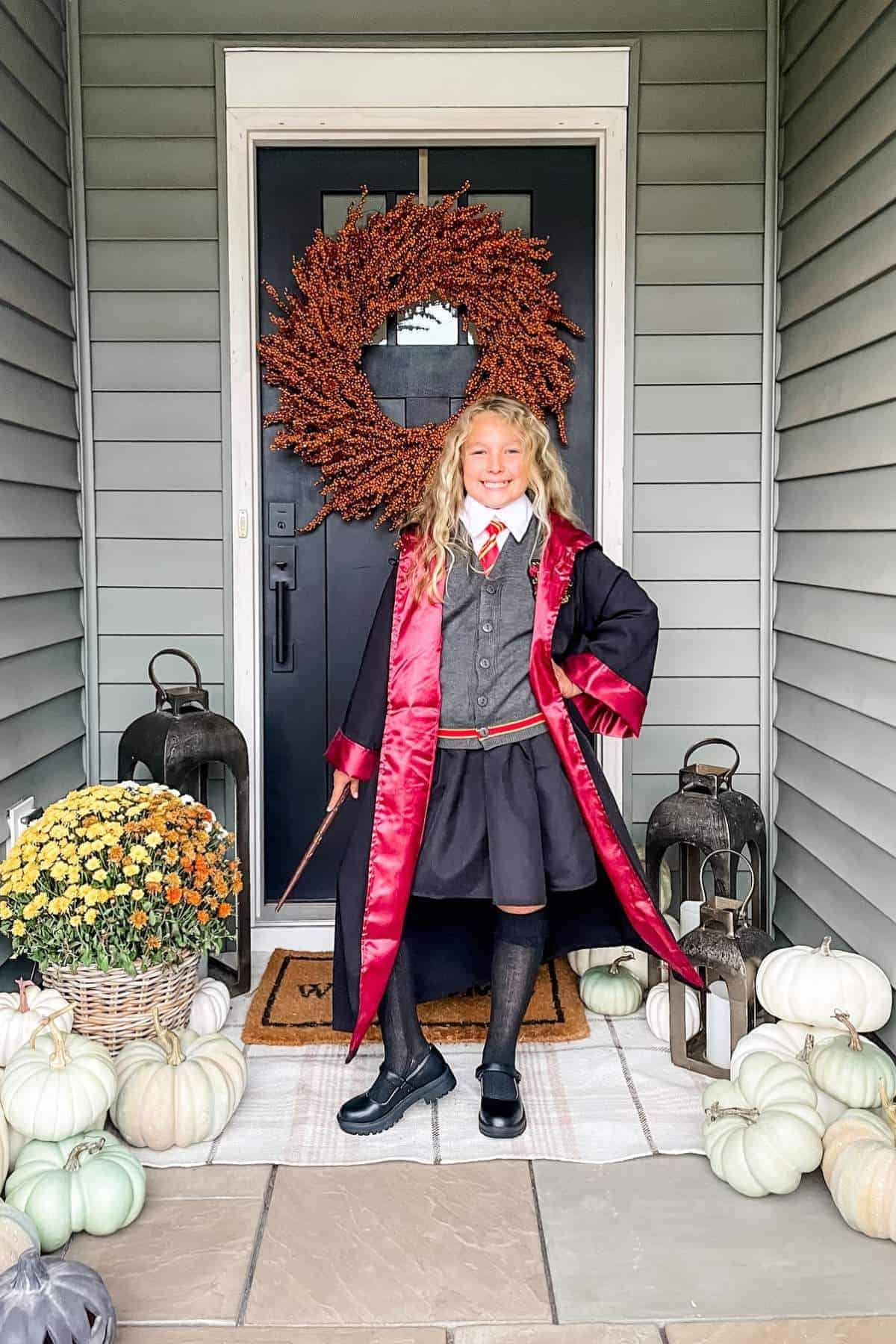 A child in a Harry Potter costume stands on a decorated porch with pumpkins, lanterns, and a fall wreath on the door.