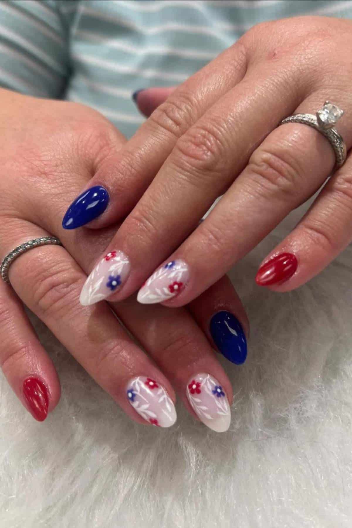 Hands with almond-shaped nails painted in red, blue, and white, with some nails featuring star accents and marble-like white designs. The person is wearing rings on both hands.