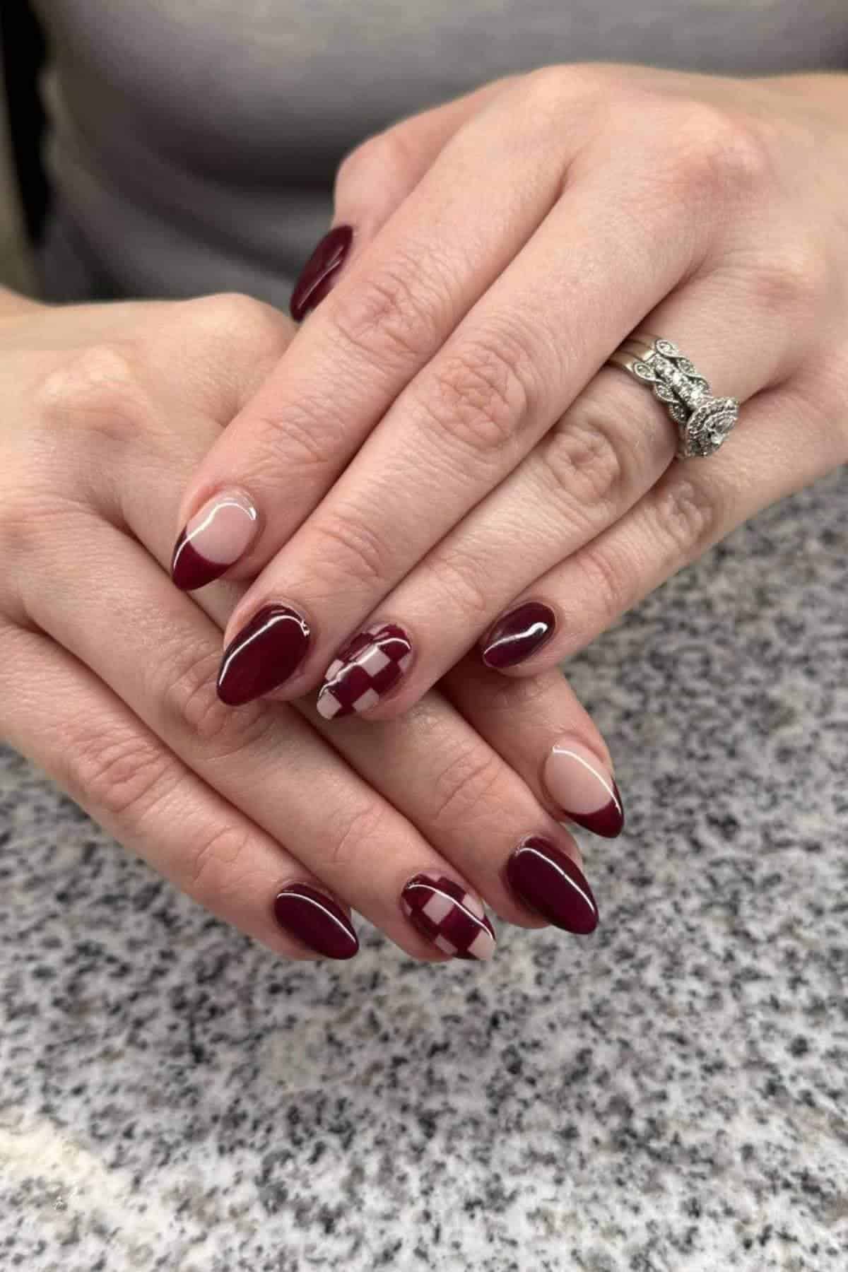 Hands with almond-shaped nails featuring a burgundy French tip design, one accent nail with a plaid pattern, and a diamond ring on the ring finger, resting on a speckled surface.