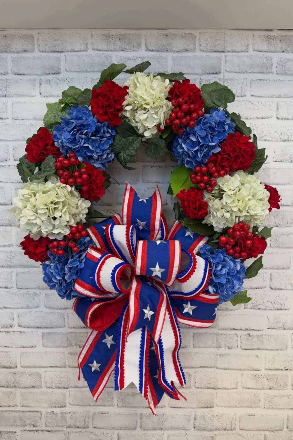 A wreath with red, white, and blue flowers, green leaves, and a large patriotic ribbon bow, displayed against a white brick wall.