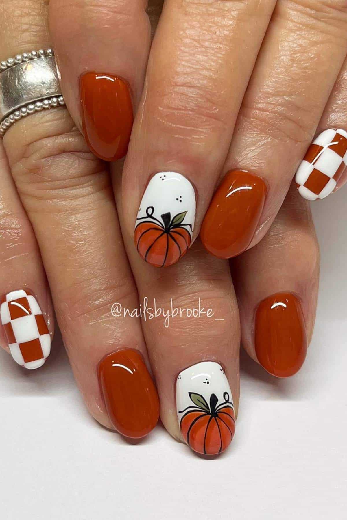 A hand with short, neatly manicured nails painted orange, with accent nails featuring pumpkins and orange-and-white checkerboard patterns.
