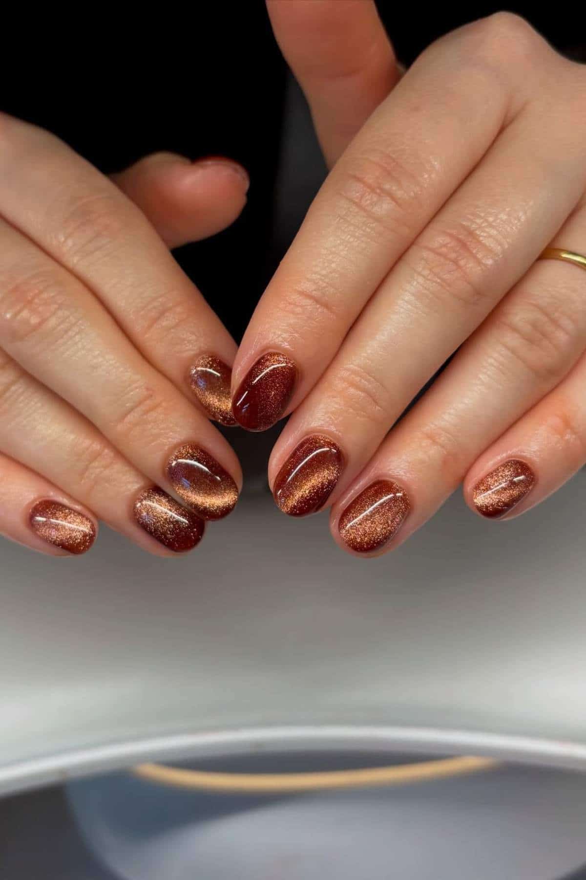Close-up of hands with short, oval-shaped nails painted in a metallic brown and gold gradient design with a glossy finish.