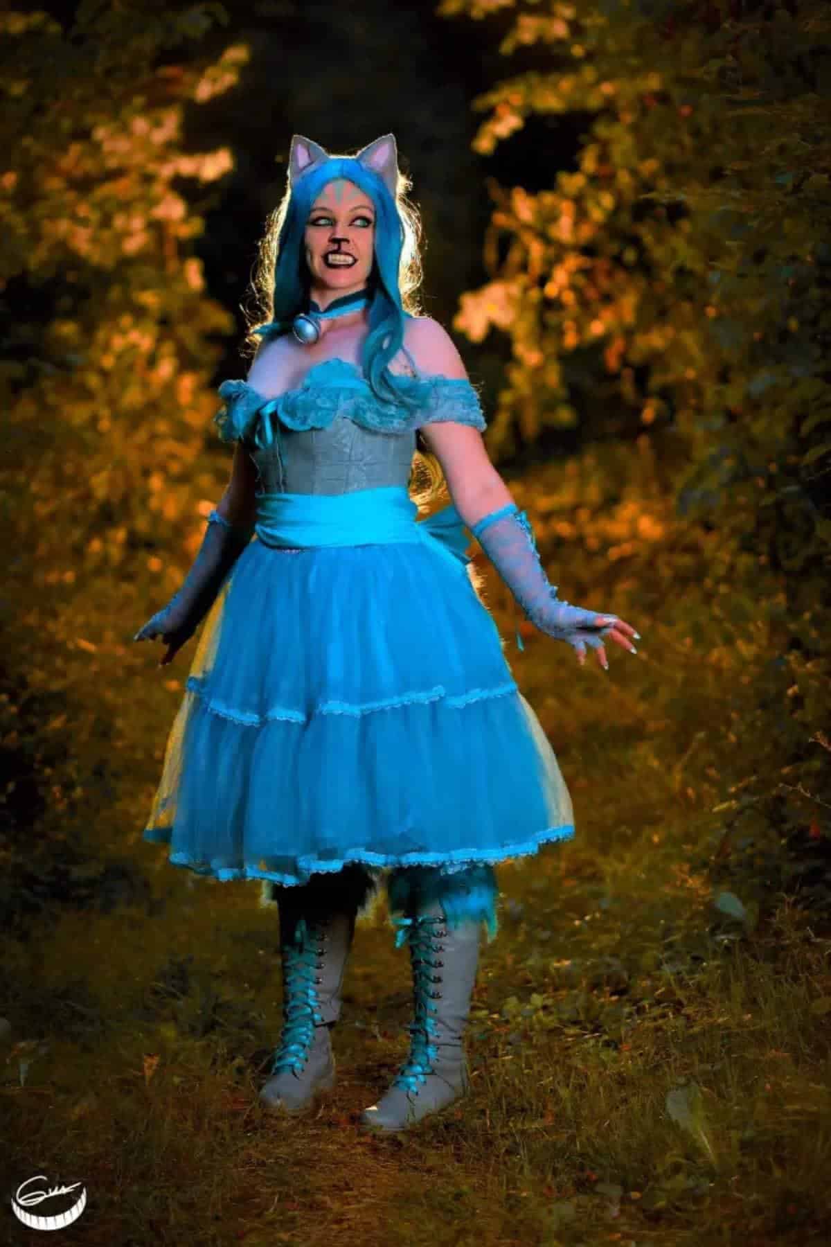 Person in a blue dress, blue wig, cat ears, and gloves poses on a path surrounded by foliage, with dramatic lighting highlighting the scene.