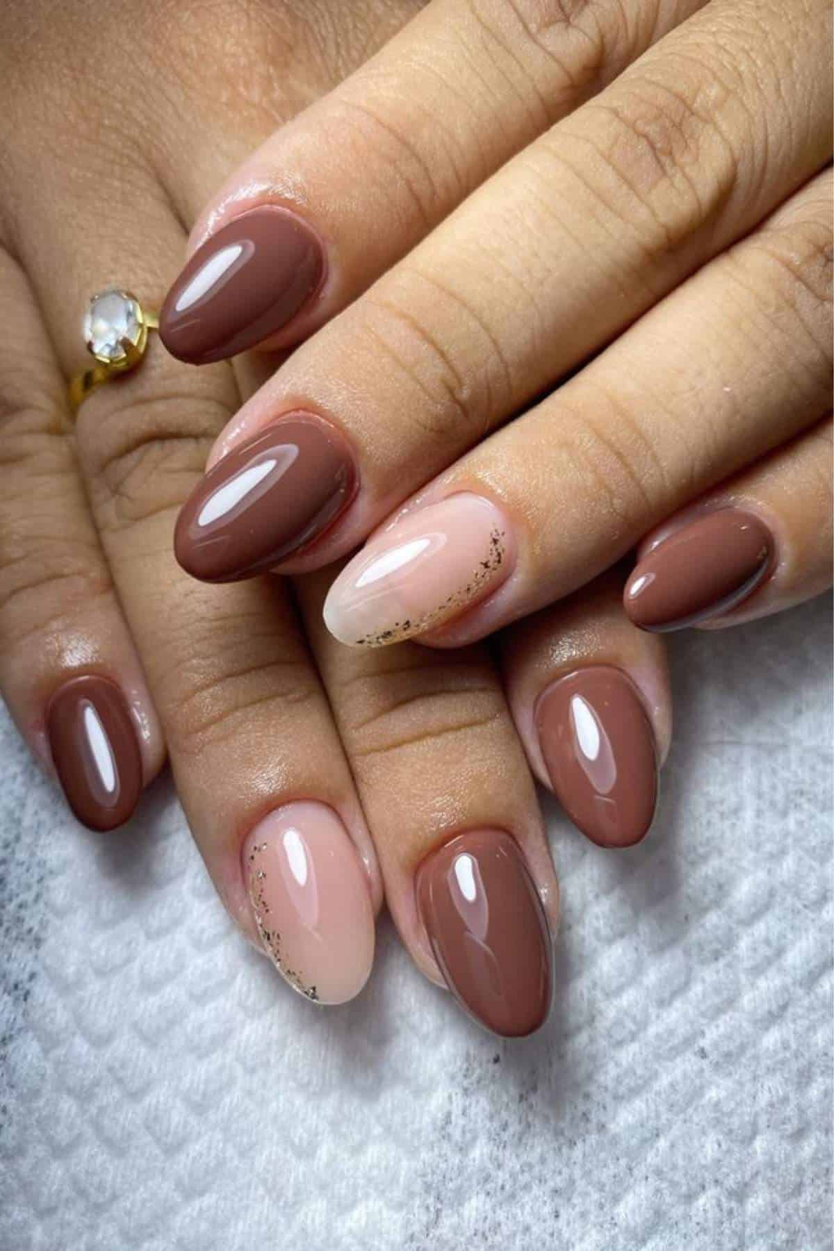 Close-up of hands with almond-shaped nails painted in glossy shades of brown and nude, with subtle gold accents on two nails.