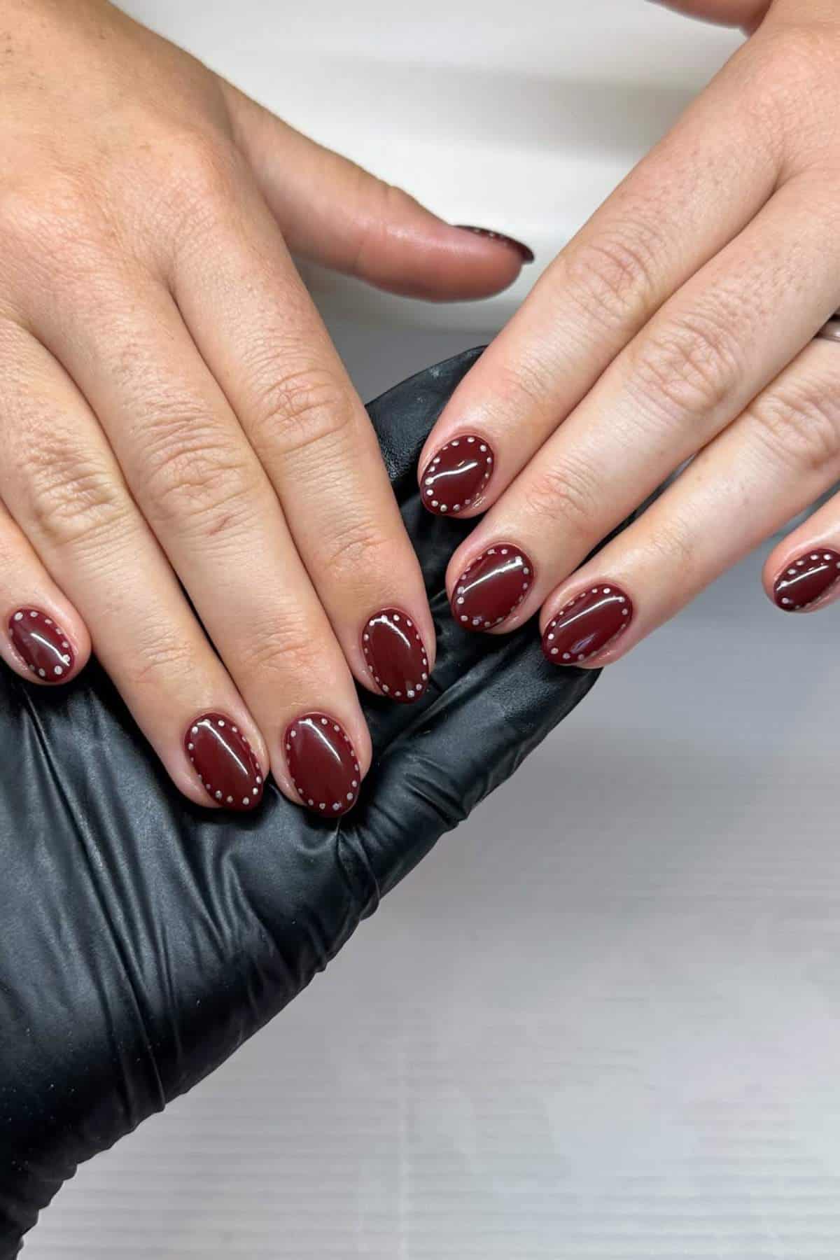 Two hands with short, oval-shaped nails painted dark red, each nail outlined with small white dots. A gloved hand supports one of the hands.