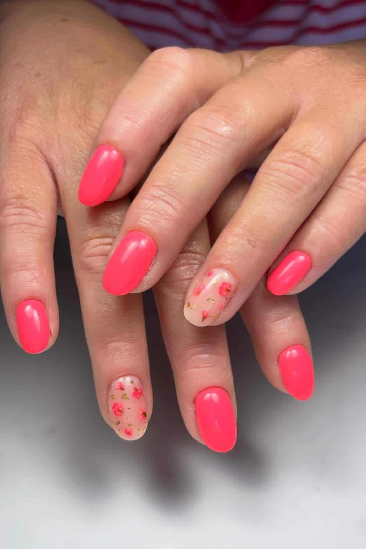 Hands with manicured nails; most nails are painted bright pink, while two accent nails have a nude base with small pink and gold floral designs.