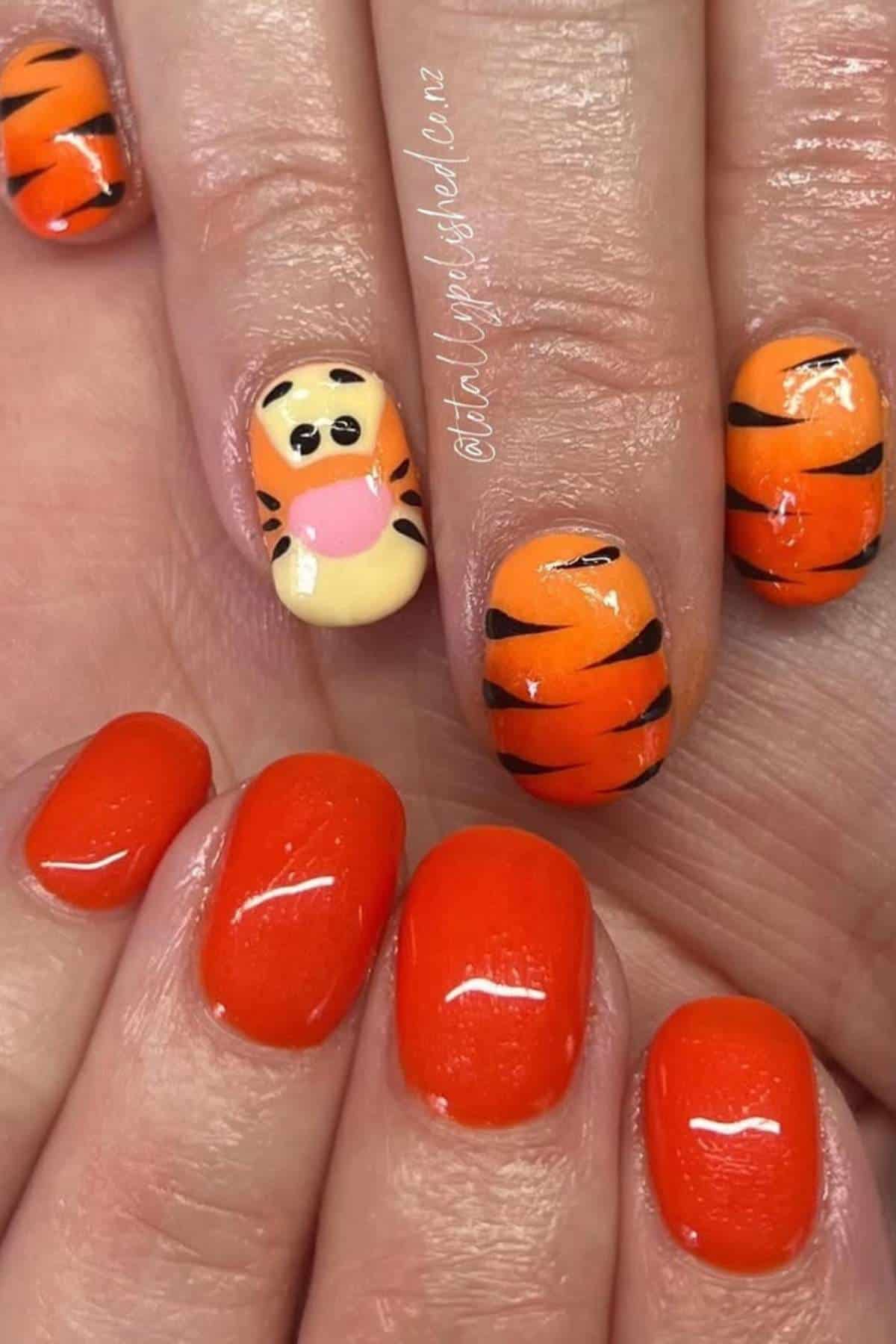 Close-up of manicured nails painted orange; some have black tiger stripes, and one nail features a cartoon tiger face with a pink tongue.