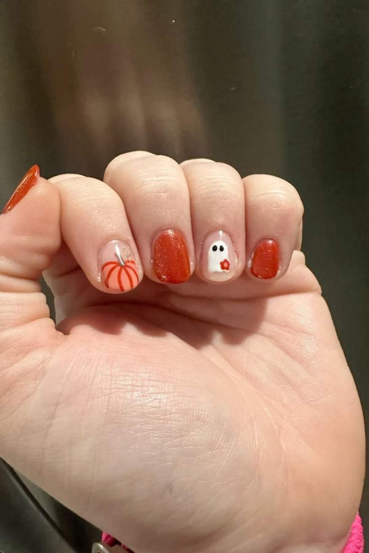 A hand with orange and white Halloween-themed nail art, featuring a pumpkin design on the pinky and a ghost on the middle finger.