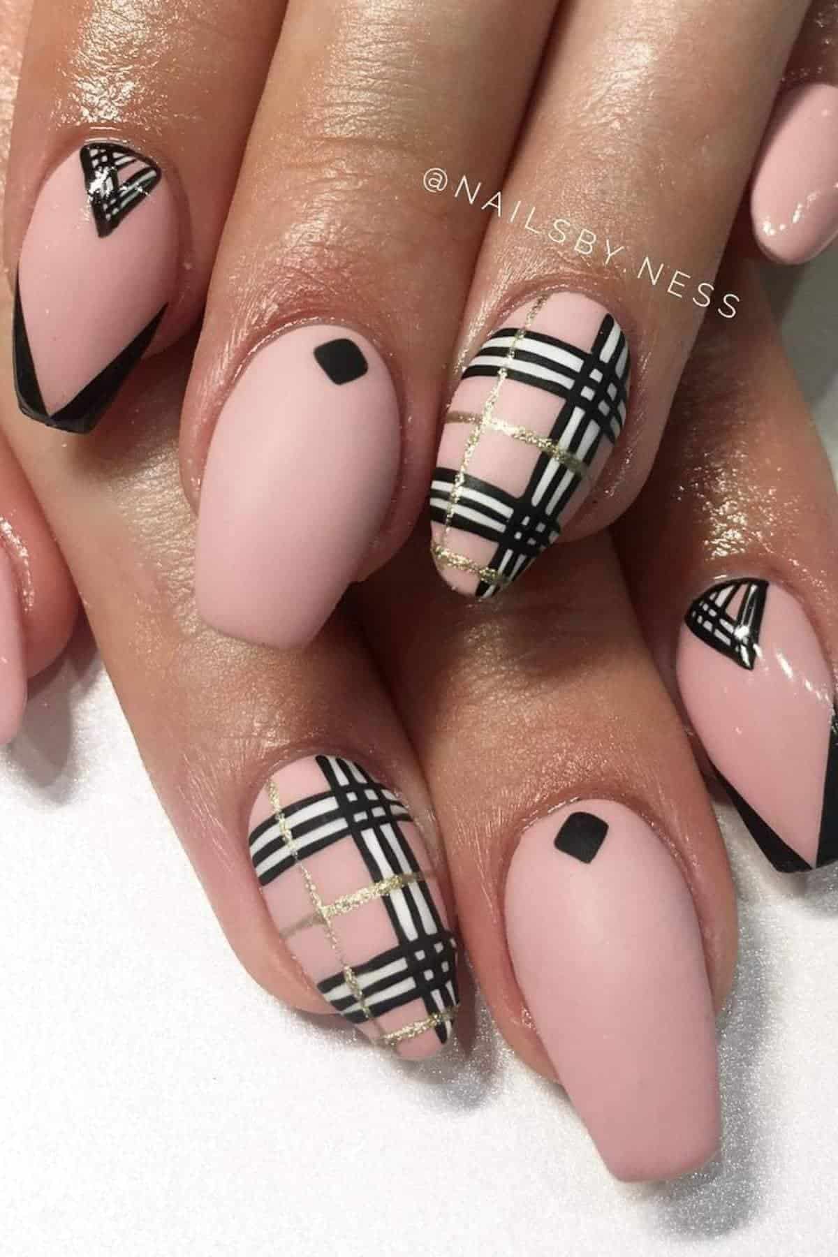 Close-up of hands with nude pink nails featuring black geometric and plaid designs, accented with gold glitter lines.