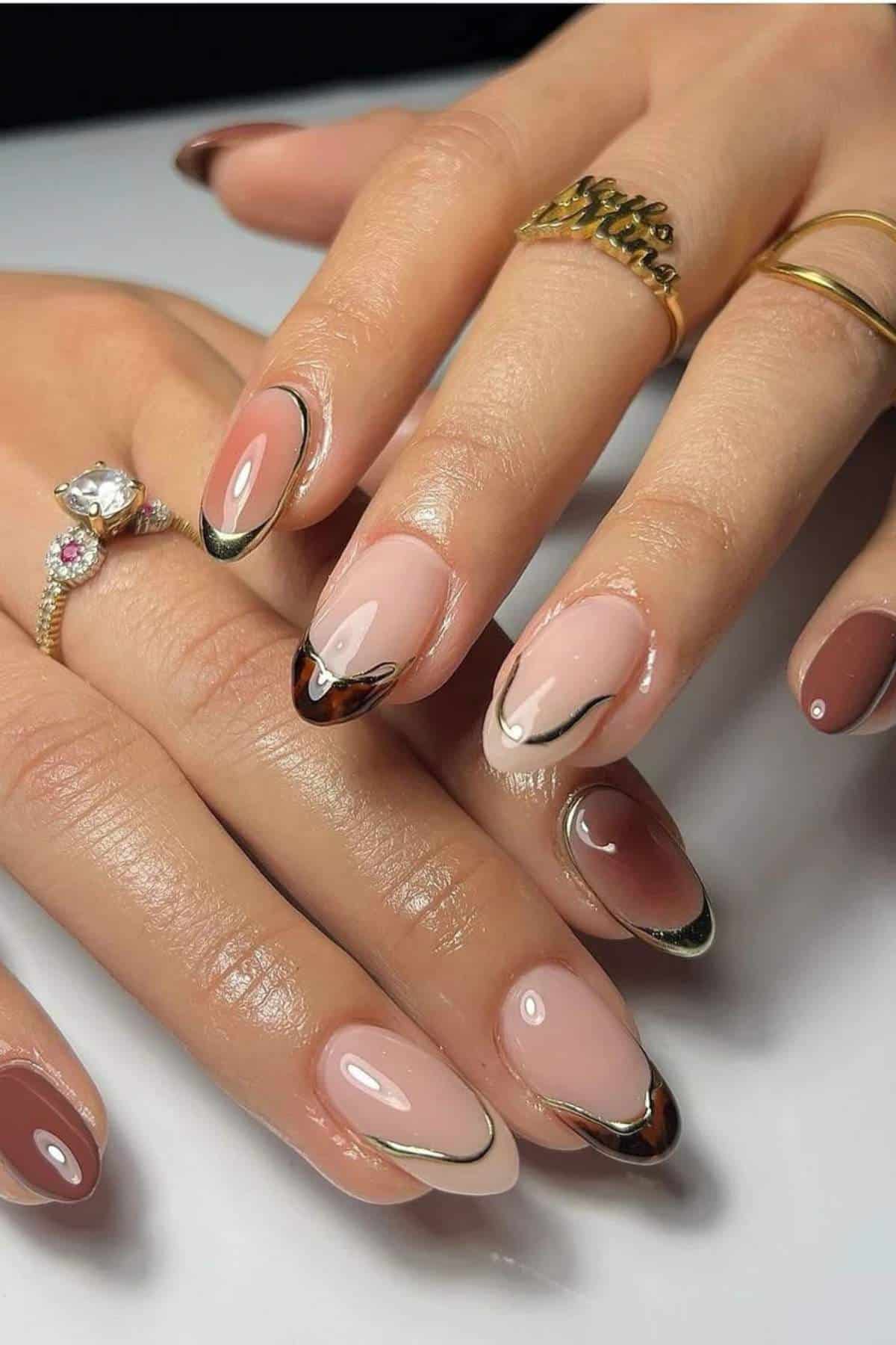 A close-up of hands with almond-shaped nails featuring a modern French manicure in nude, brown, and beige tones with metallic accents.