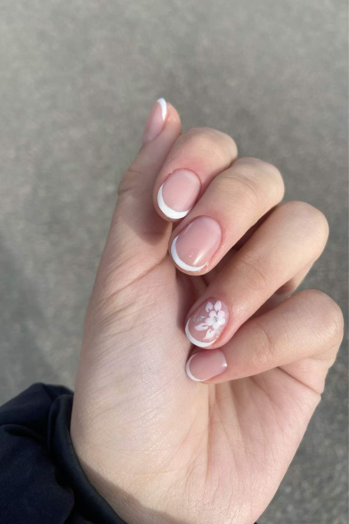 A hand with short, natural-shaped nails featuring a French manicure and a white floral design on the ring finger.