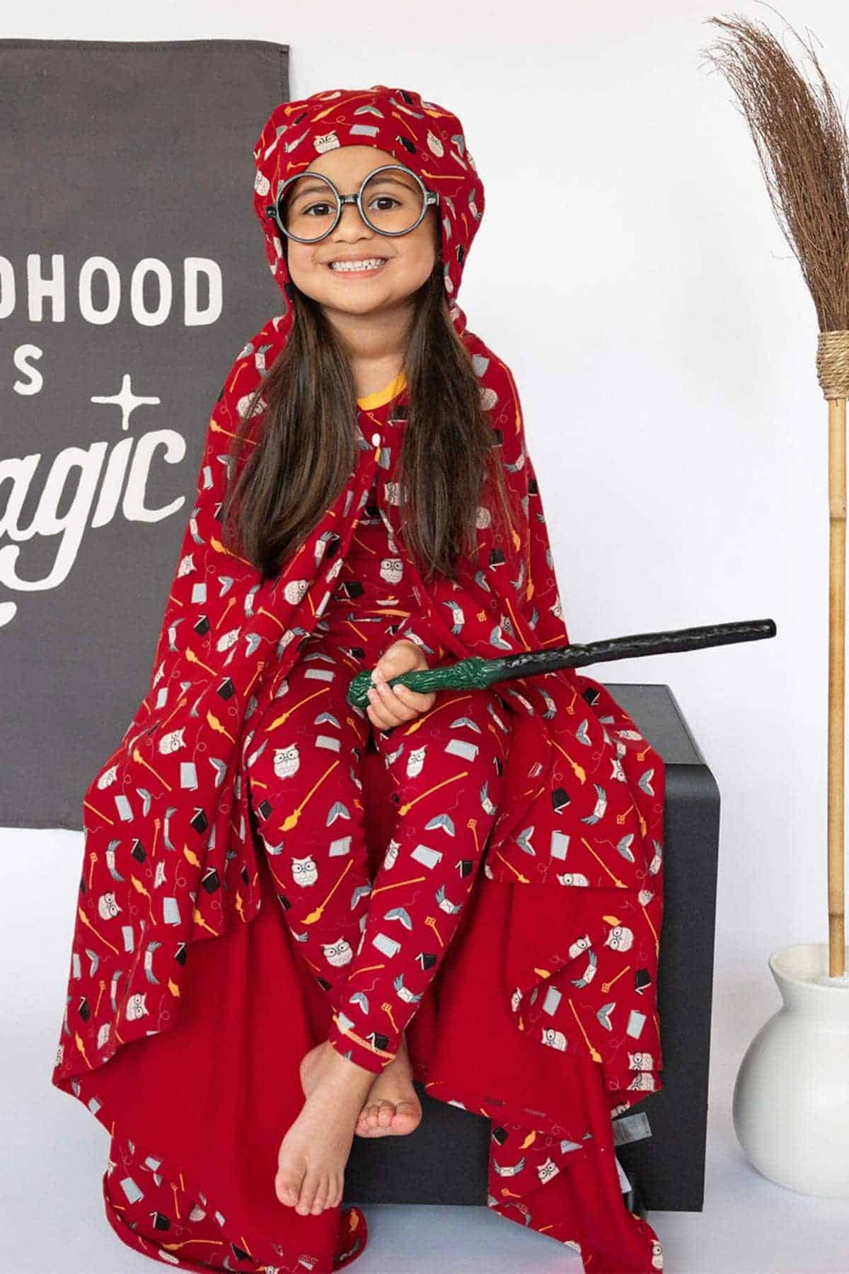 A young girl in red pajamas with wizard-themed prints, wearing round glasses and a matching headwrap, sits barefoot holding a wand, next to a broom and a sign that reads “Childhood is magic.”.