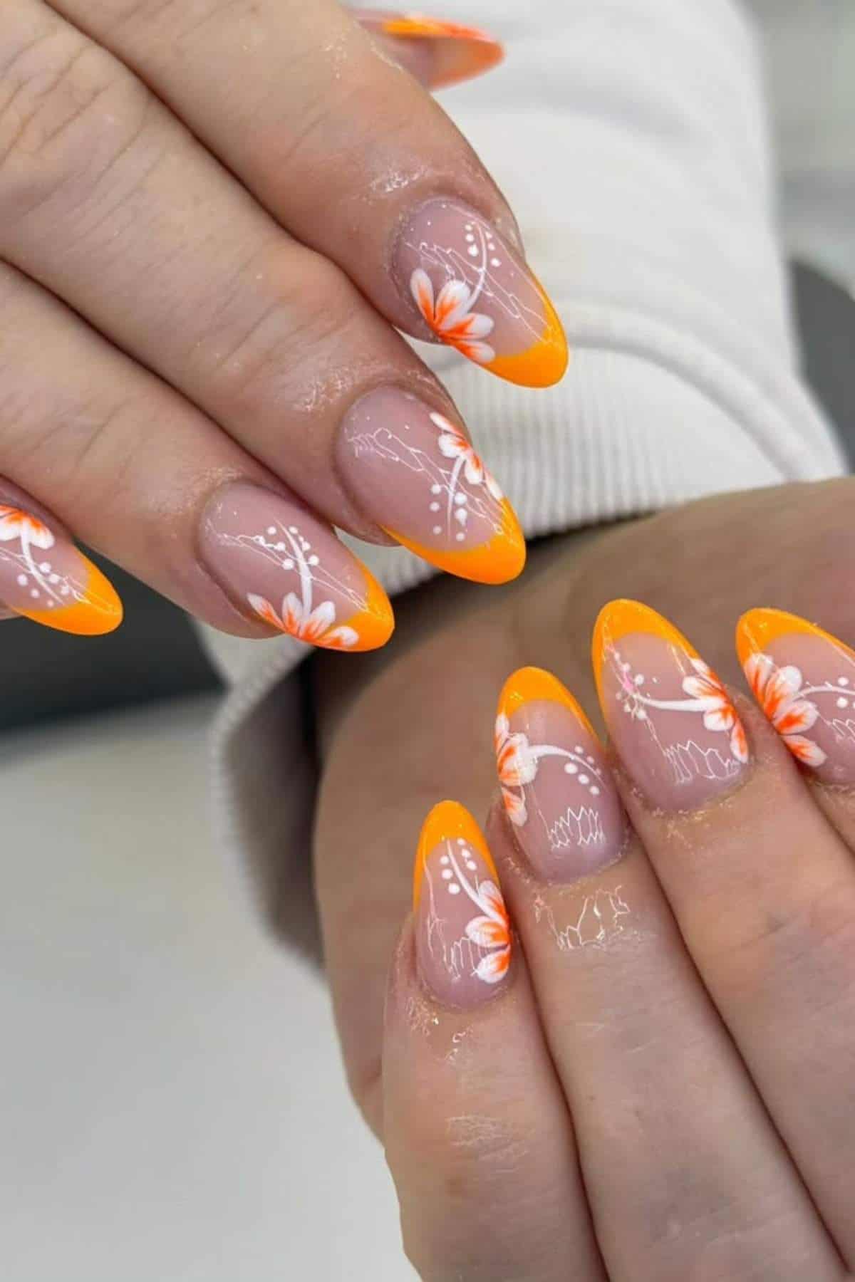 Hands with almond-shaped nails featuring orange French tips and white floral designs on a nude base.