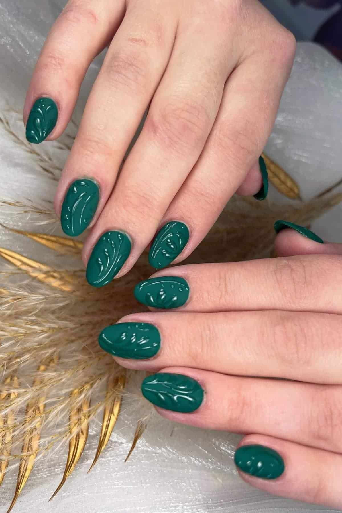 Hands with green, almond-shaped nails featuring a raised, textured leaf-like design, resting on a light fabric with dried wheat stalks.