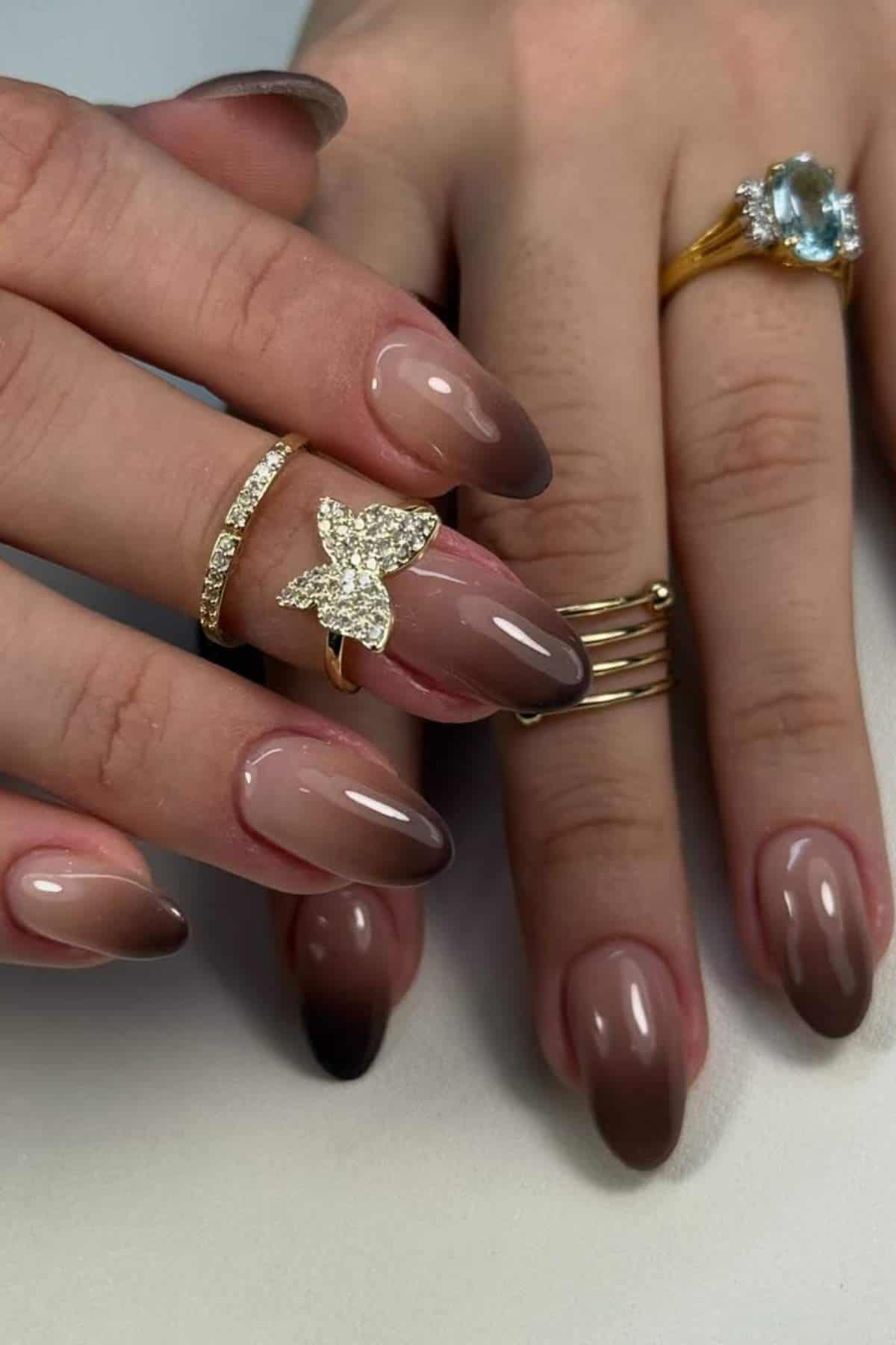 Close-up of hands with almond-shaped, brown ombré nails, wearing gold rings, including one with a gemstone and another with a decorative butterfly design.