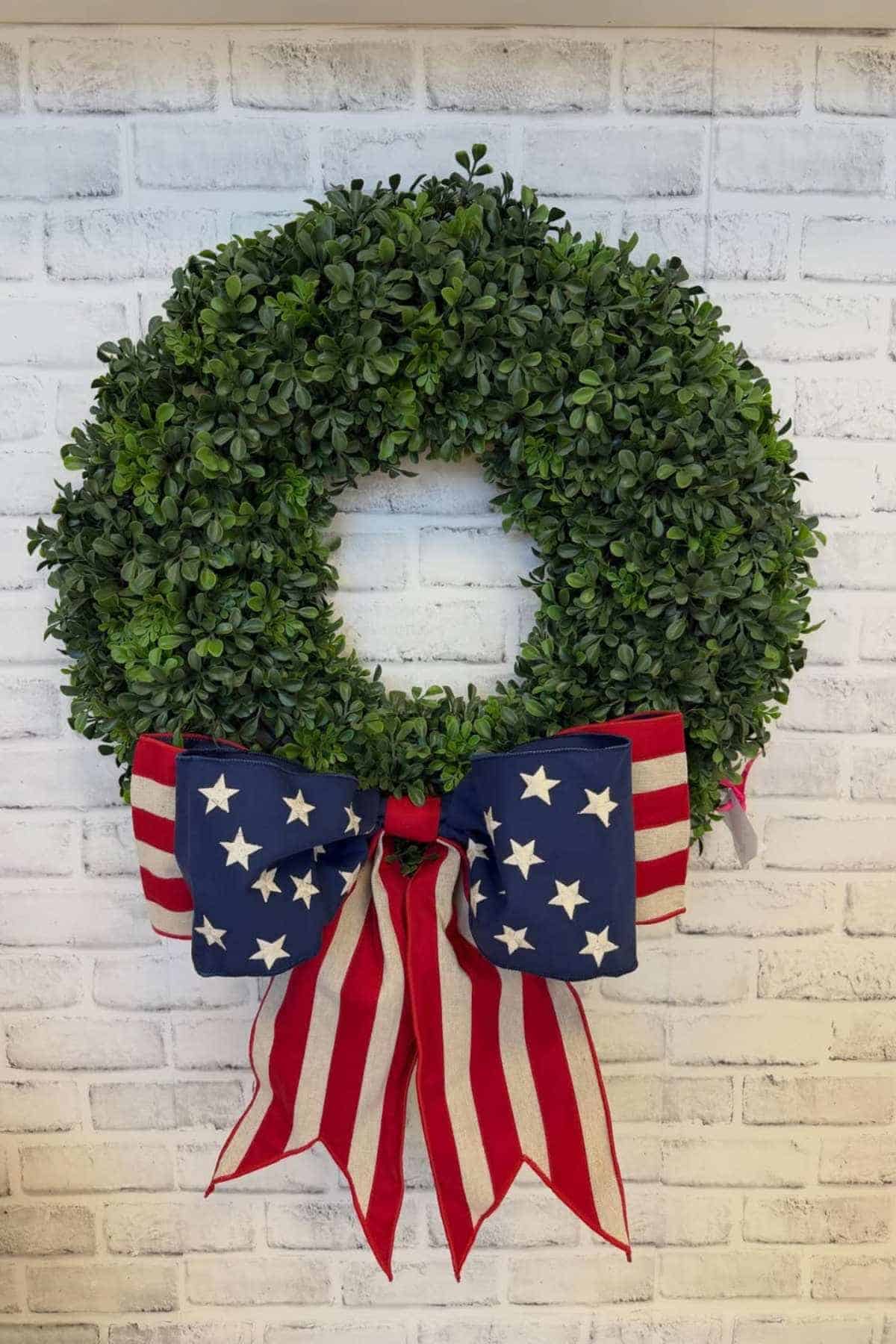 A green wreath with a large bow featuring the stars and stripes of the American flag is hanging on a white brick wall.