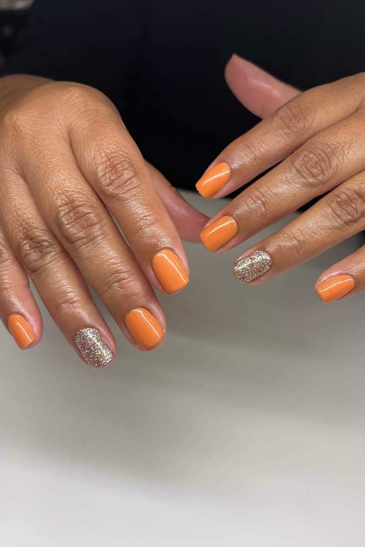 Hands with short, neatly manicured nails painted orange; one nail on each hand features a silver glitter accent.