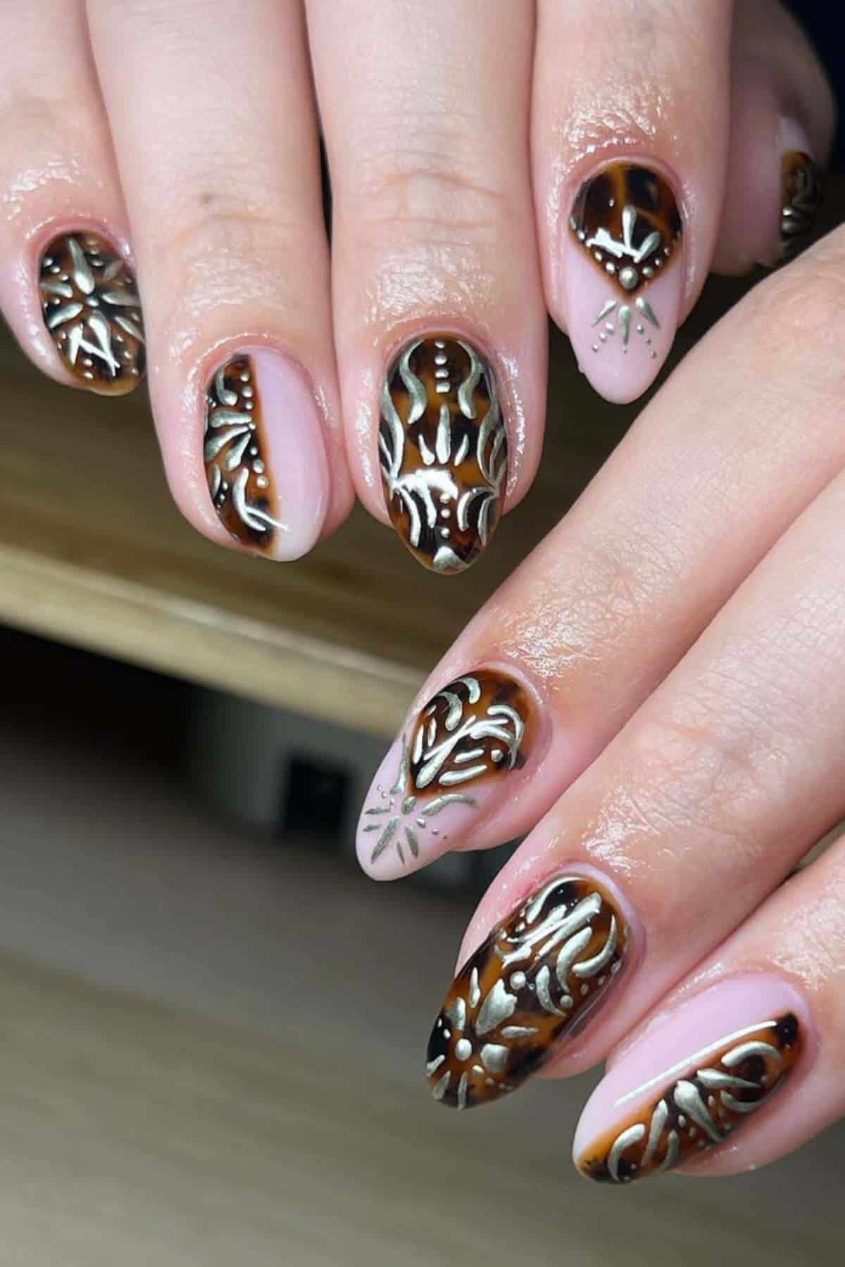 Hands with almond-shaped nails featuring intricate brown, white, and silver geometric and floral patterns on a nude base.