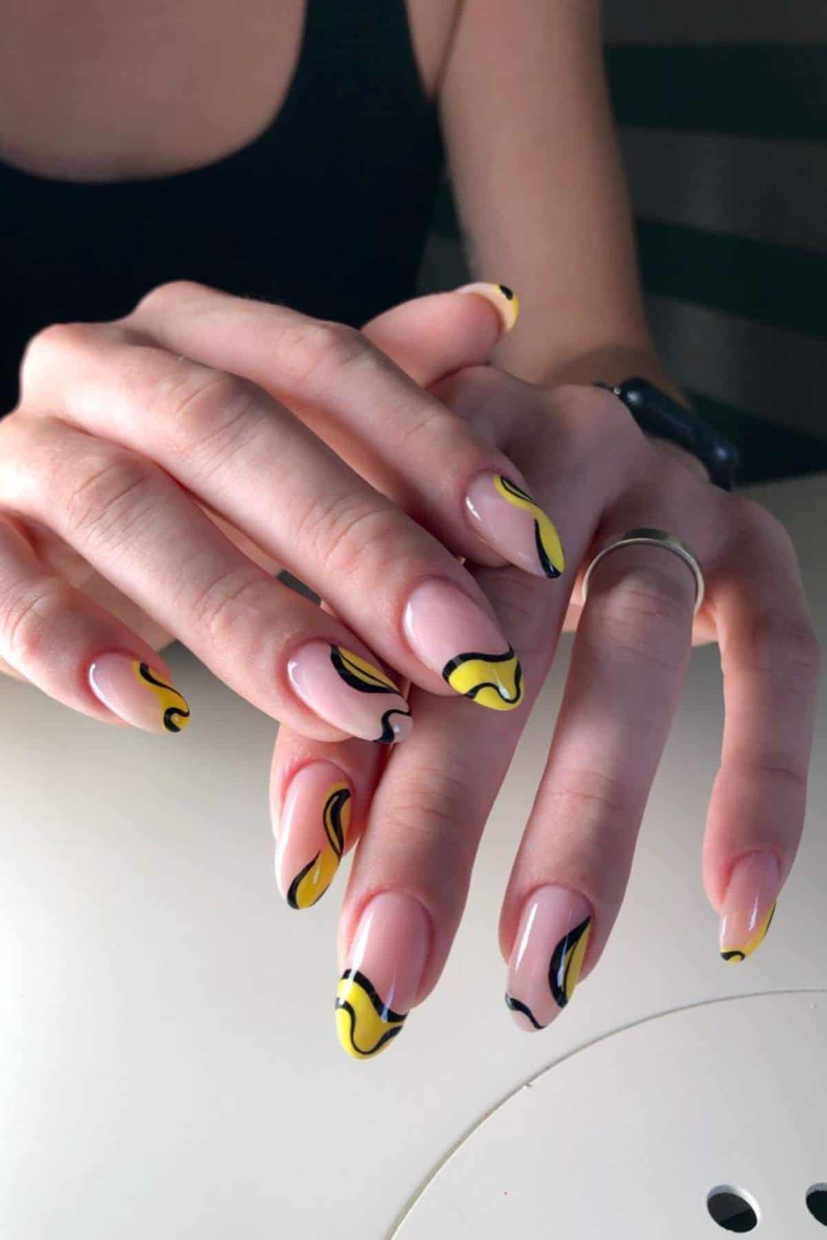 A person with almond-shaped nails featuring a nude base and abstract yellow and black swirl designs on each nail.