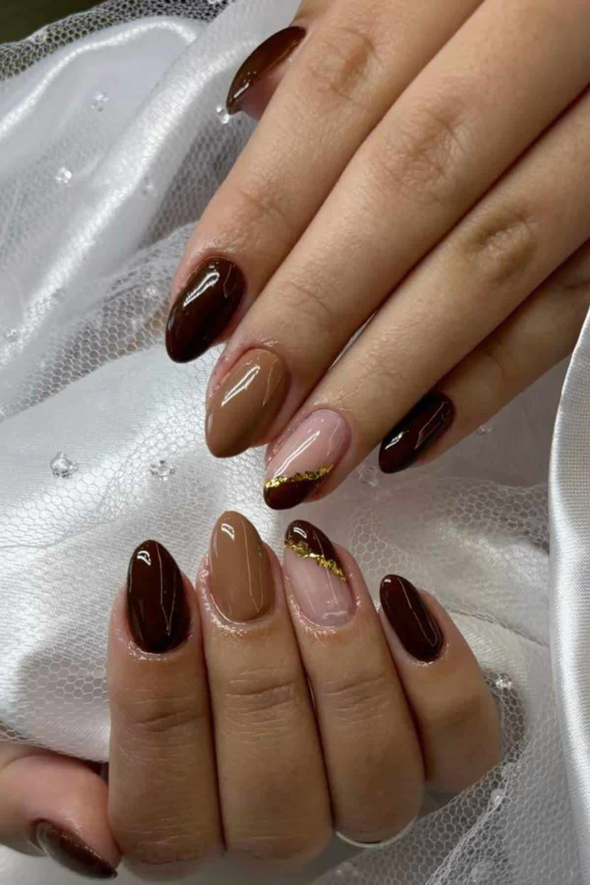 Hands with almond-shaped nails painted in shades of brown and nude, featuring gold accents and a glossy finish, resting on white fabric.