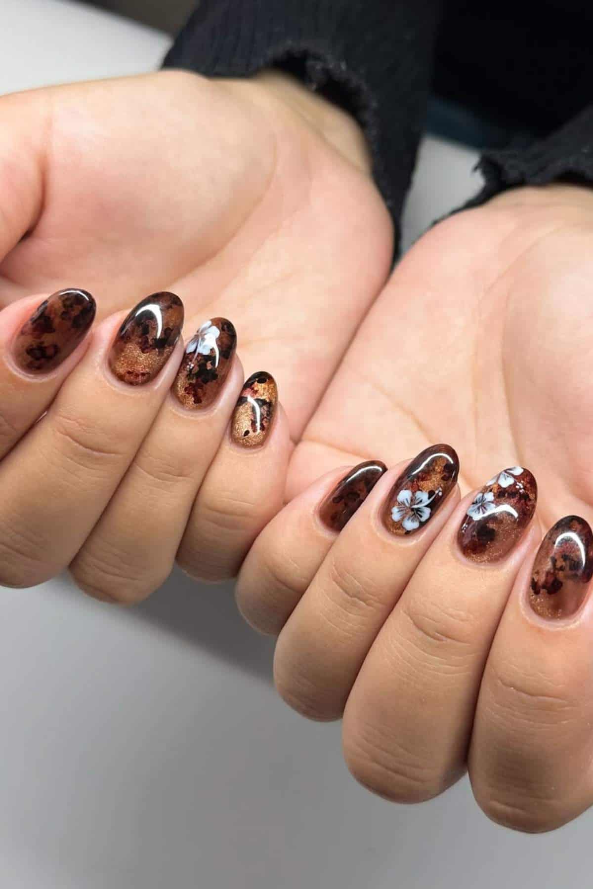 Hands with oval-shaped nails featuring a brown marble design and white floral accents on a few nails, against a plain background.